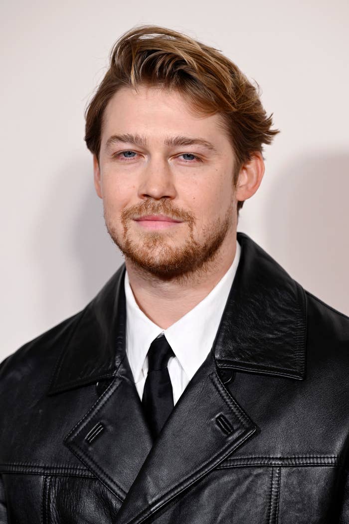 Joe Alwyn wears a black leather coat, white shirt, and black tie