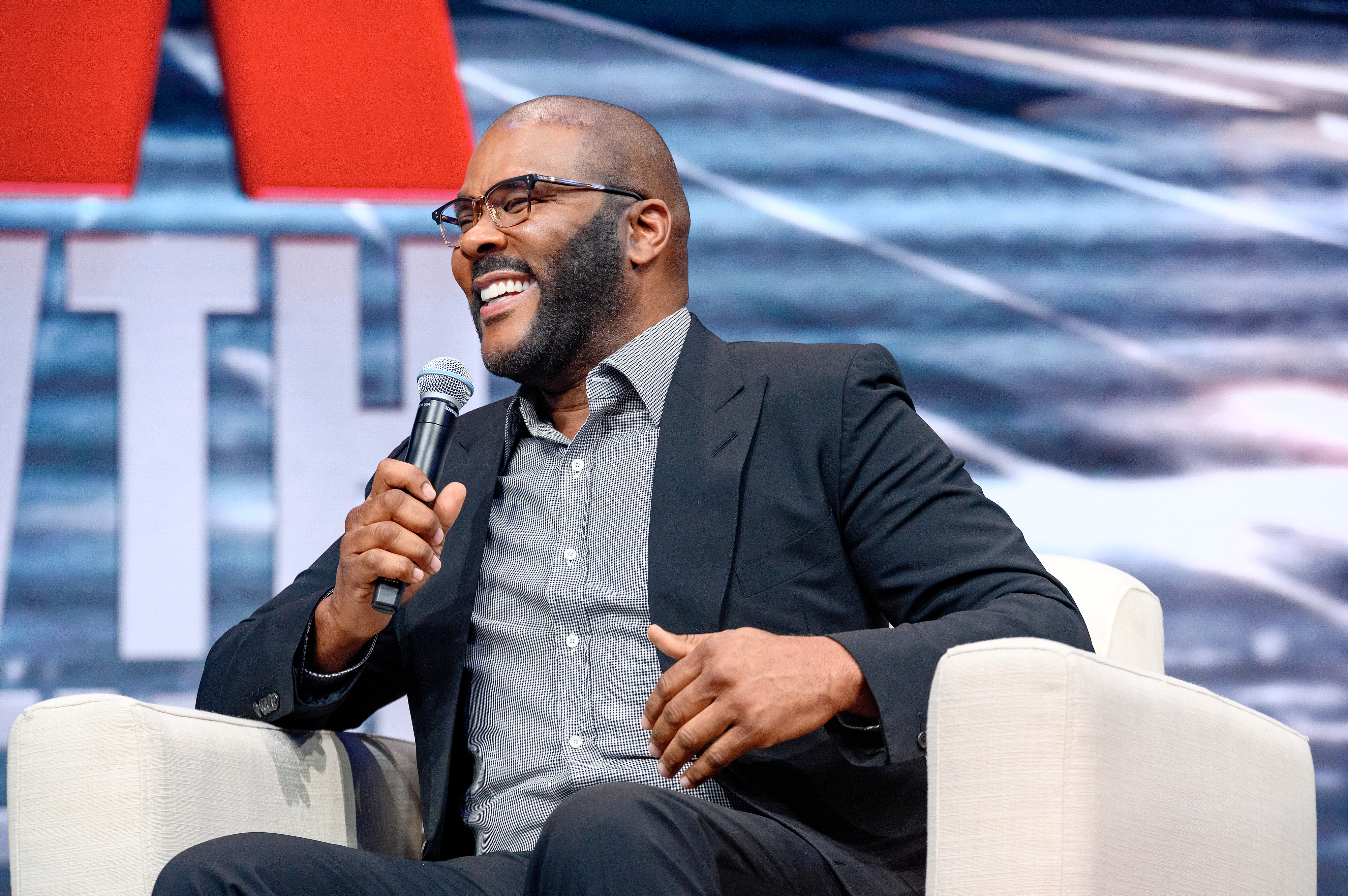 Tyler Perry, in a suit and open-collar shirt, smiling while holding a microphone and sitting in a chair during an event with &quot;TYTH&quot; partially visible in the background
