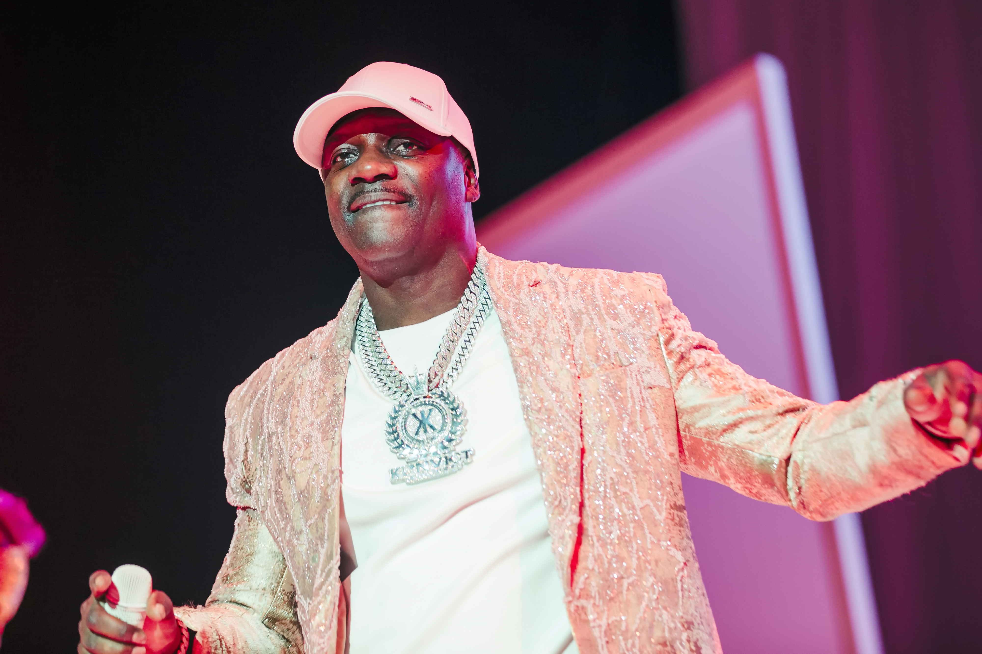 Akon on stage wearing a white hat, a textured blazer, a white T-shirt, and a large chain necklace