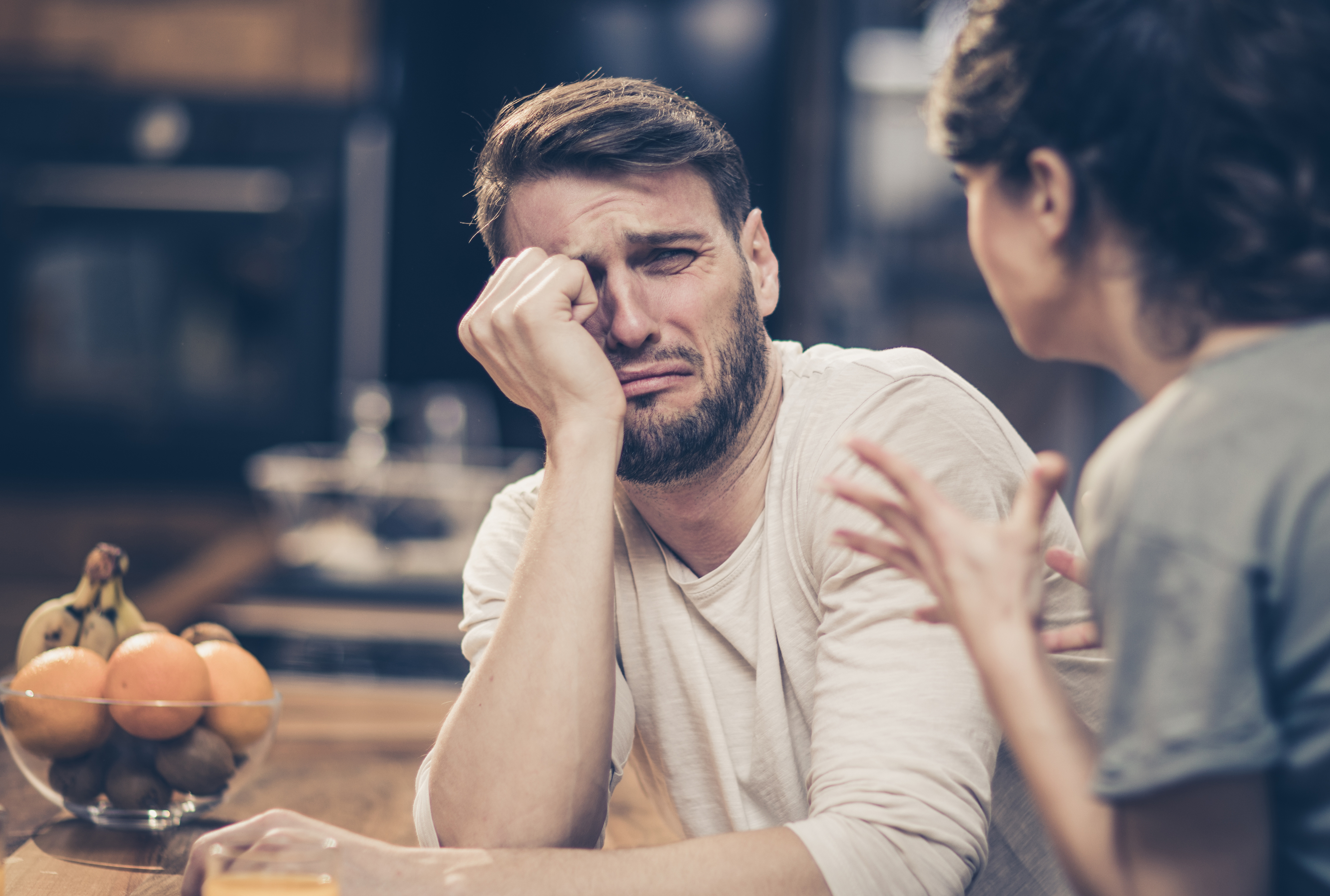 A man, looking distressed, holds his head and appears to be crying at a kitchen table with a bowl of fruit. A woman next to him seems to be speaking