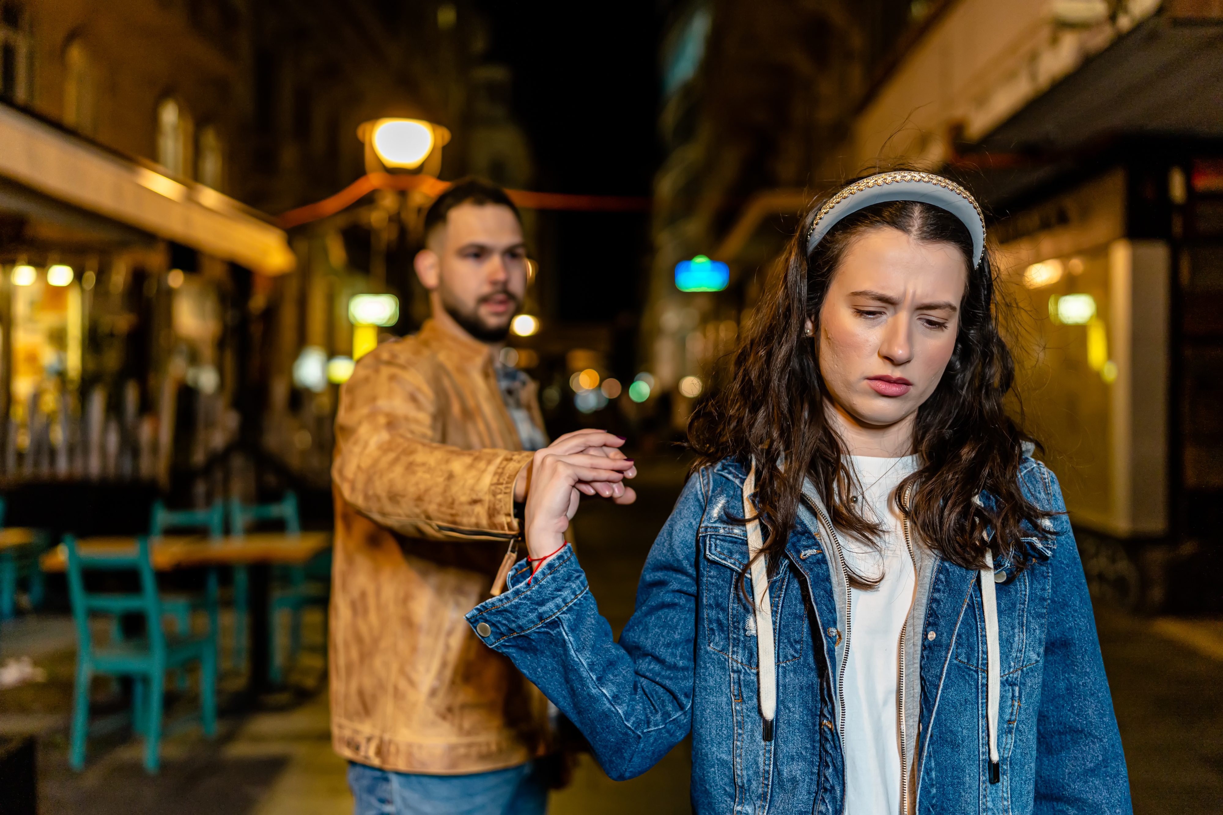 A man and a woman outside at night. The man is reaching for the woman's hand, while the woman looks upset and pulls away