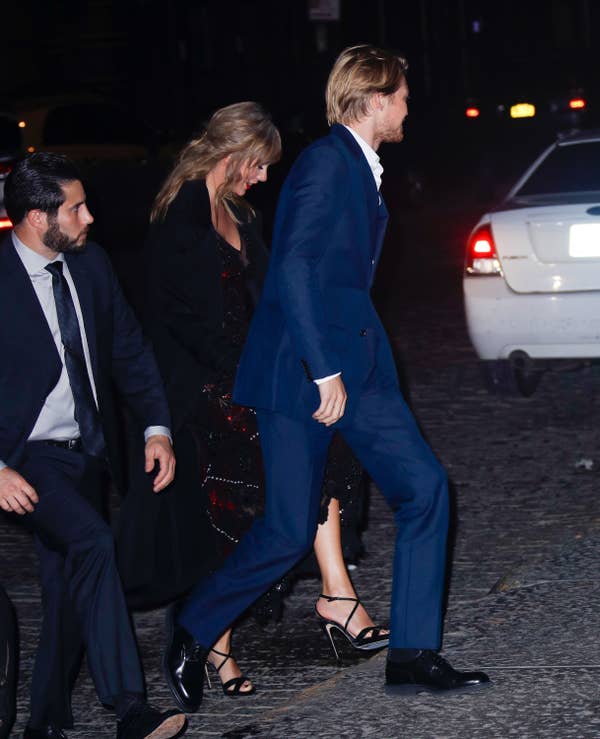 Joe Alwyn and Taylor Swift are pictured walking down a street at night, accompanied by a man in a black suit. Joe wears a blue suit, and Taylor wears a black dress