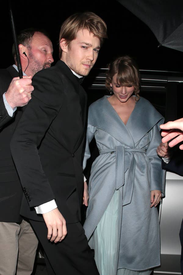 Joe Alwyn in a black suit and Taylor Swift in an elegant coat with a dress underneath leaving an event, escorted by security personnel