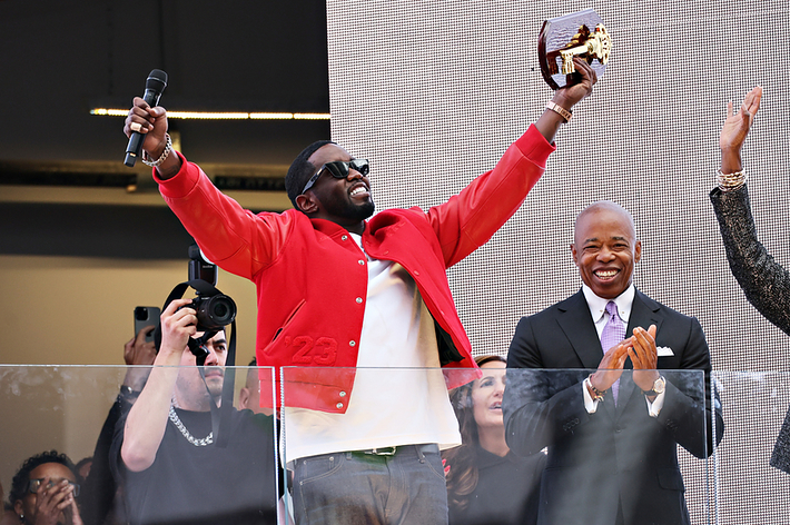 Person in a red jacket holds an award and cheers on stage. Another person in a suit stands next to him, applauding. People in the audience also cheer and clap