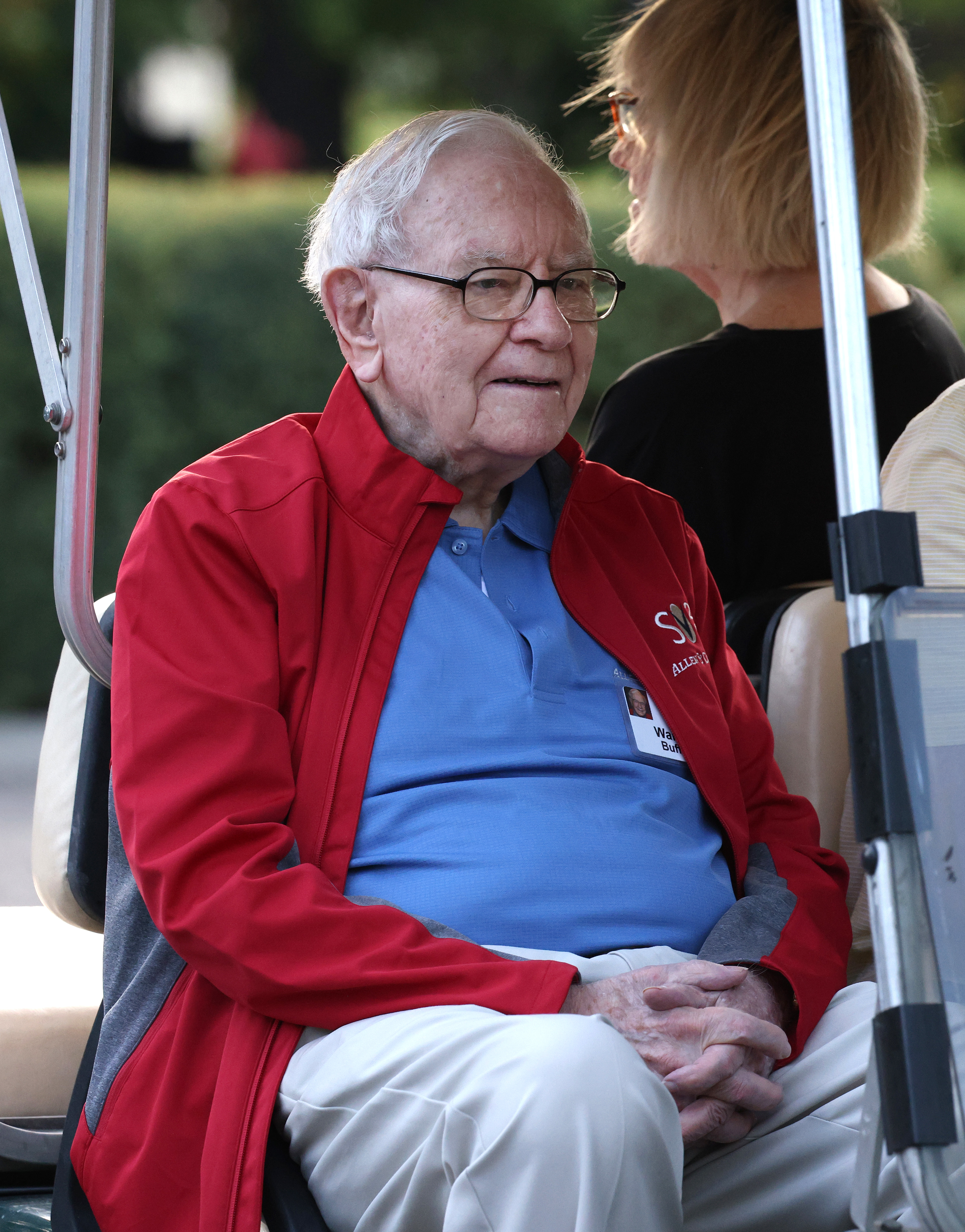 Warren Buffett sits in a golf cart wearing a red jacket, blue shirt, and beige pants, alongside a woman with short blonde hair facing away