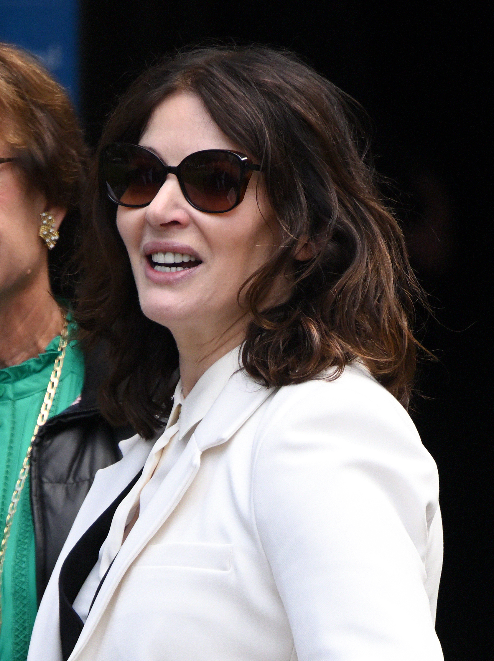 Woman in a white blazer and sunglasses smiling, with another person partially visible next to her