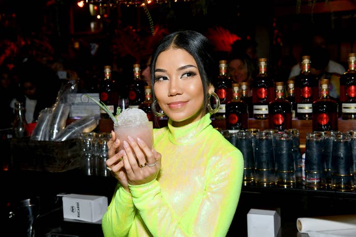 Jhené Aiko holds a cocktail while standing behind a bar with various bottles and glasses in the background. She wears a high-necked outfit