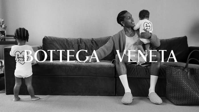 A$AP Rocky sits on a sofa holding a child wearing an &quot;I love dad&quot; onesie, with another child facing away. Bottega Veneta logo is displayed
