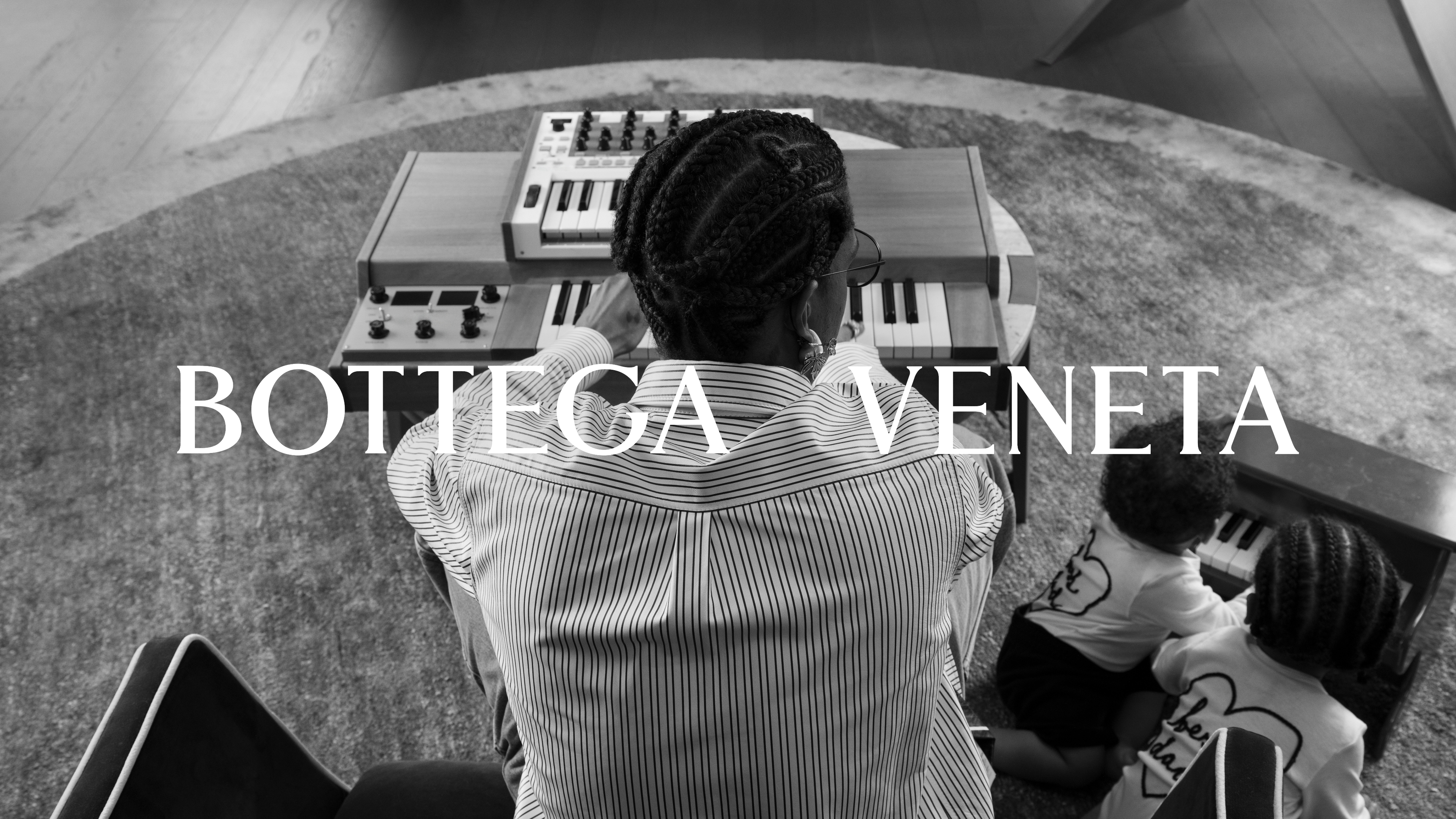 A person with braided hair and children are seated, viewed from behind, with text &#x27;Bottega Veneta&#x27; overlaying the image. A keyboard instrument is in front of them