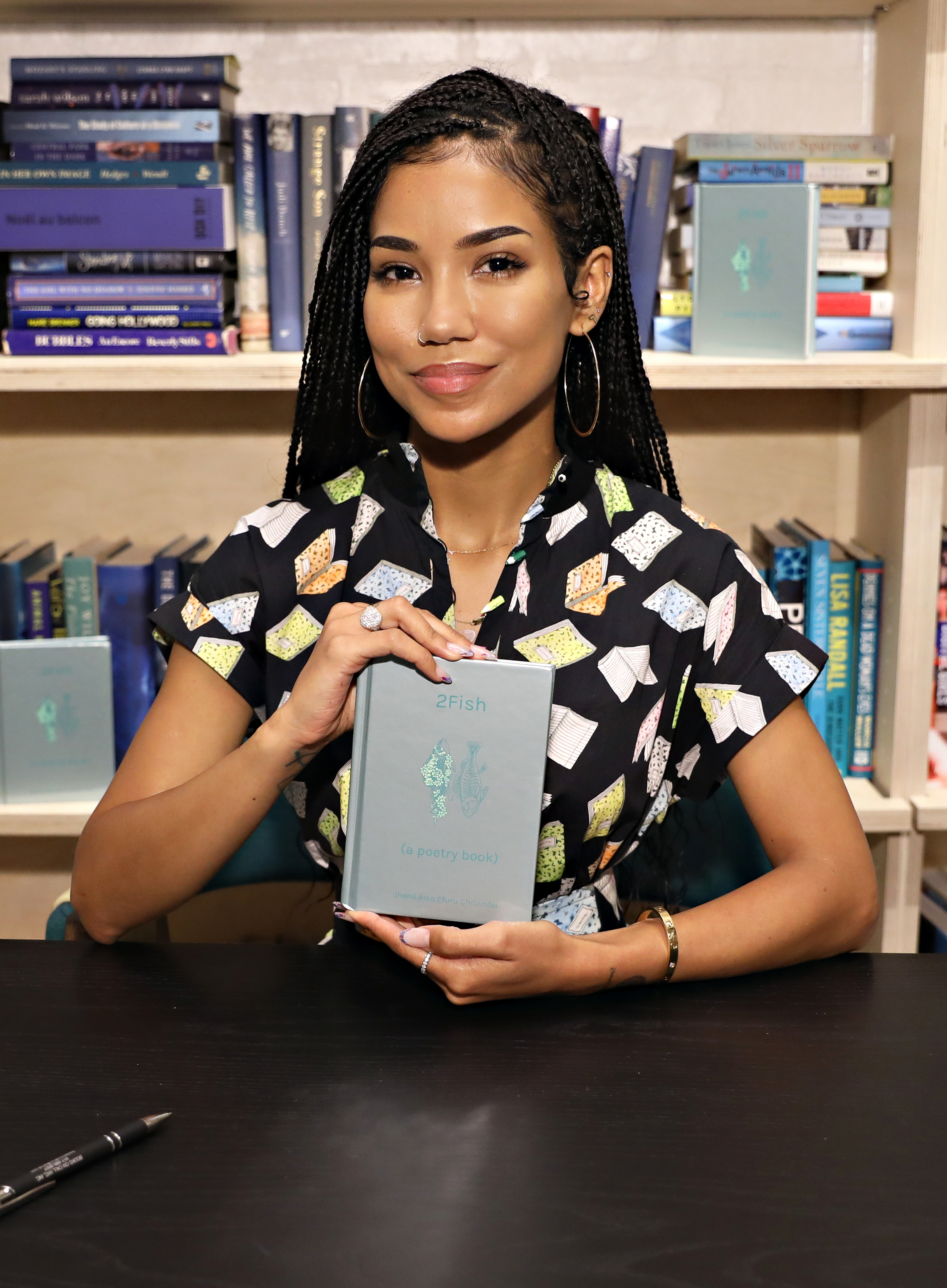 Jhené Aiko at a book signing event, holding her poetry book "2Fish" and smiling at the camera, with bookshelves in the background