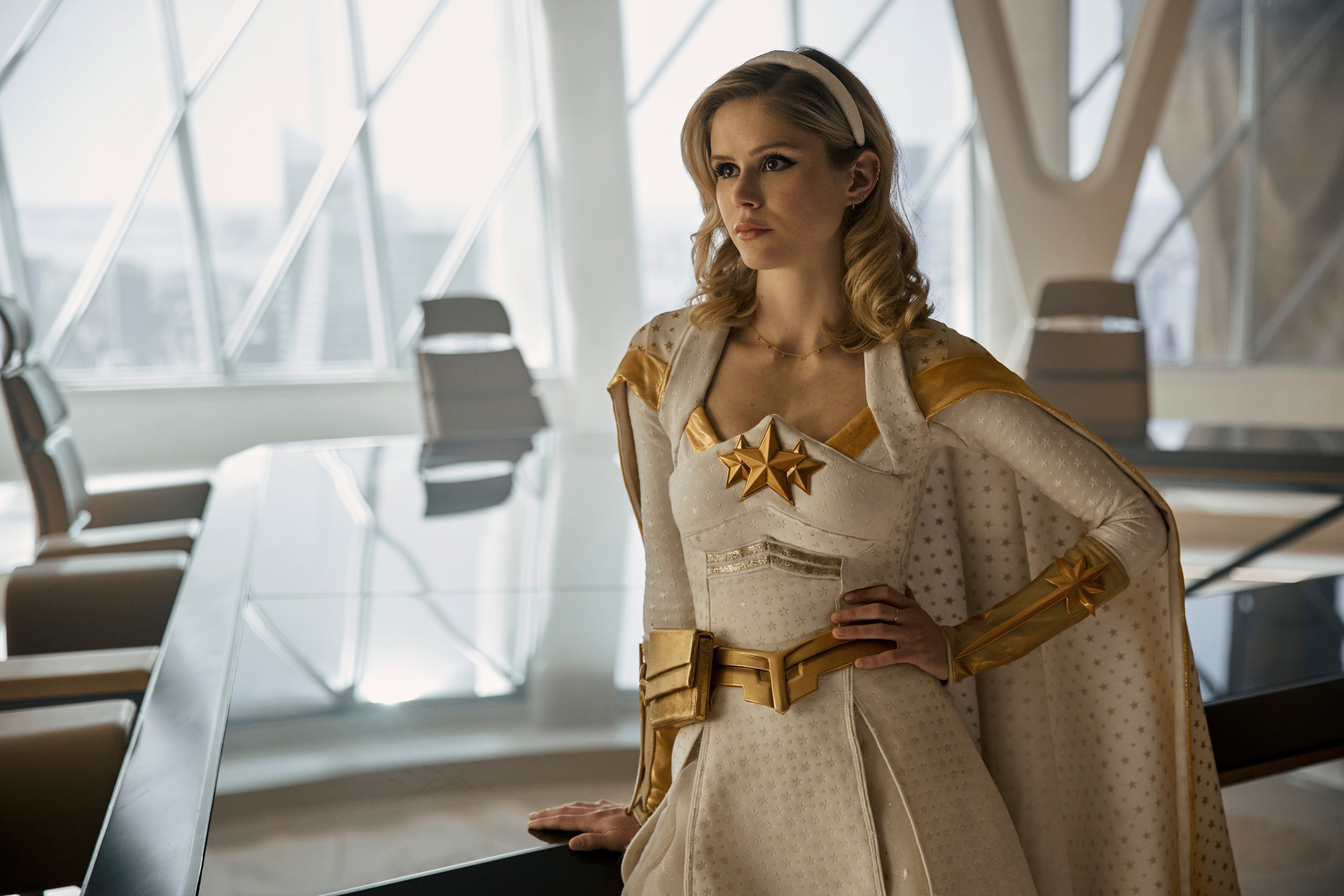 Erin Moriarty dressed as her character Starlight from "The Boys" TV series, wearing a white and gold superhero outfit with a star emblem, standing in an office setting
