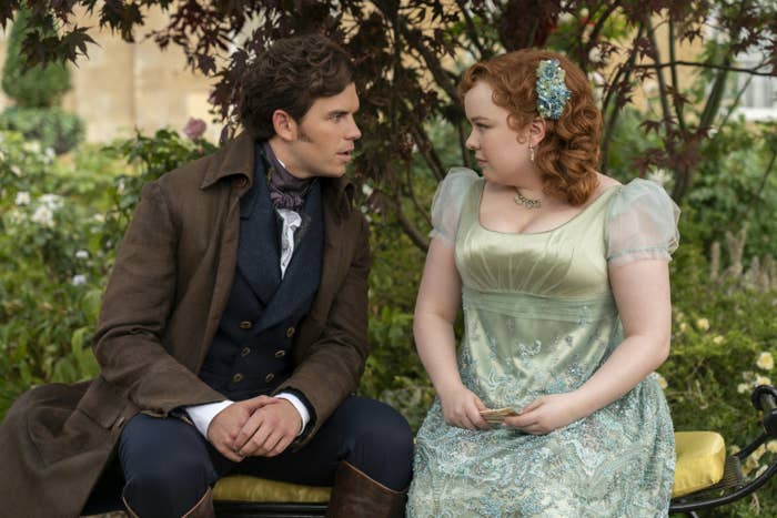 Nicola Coughlan wearing a light dress with lace detailing and florals in her hair sits on a bench with Luke Newton in period attire. They look at each other