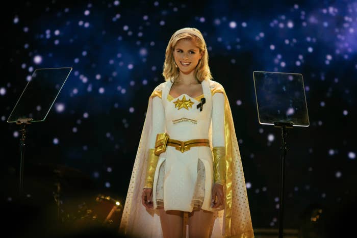 Erin Moriarty dressed as her character Starlight in a white and gold superhero costume, standing between two teleprompters with a starry background