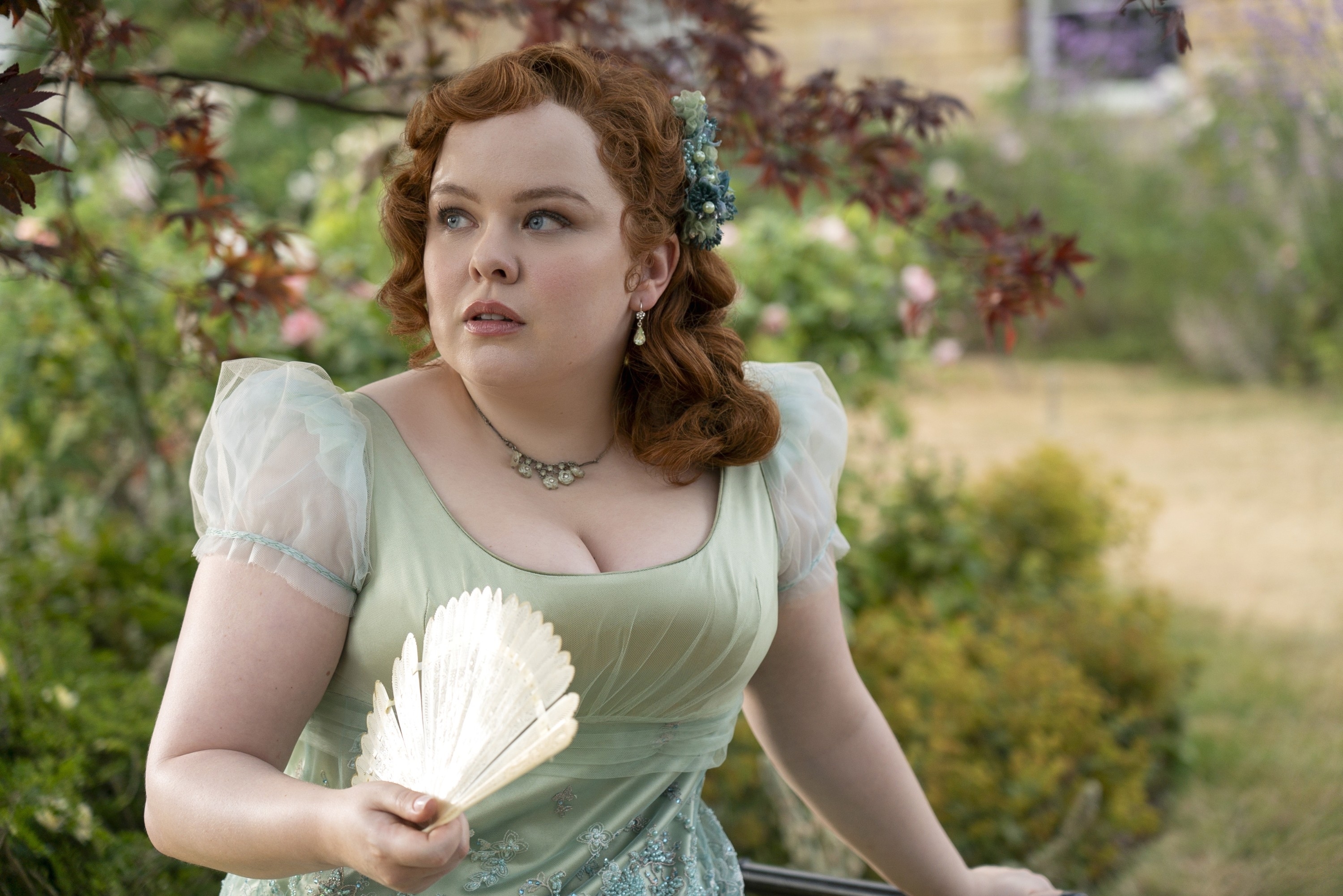Nicola Coughlan in a period costume dress, holding an open fan, looking to the side with a garden background
