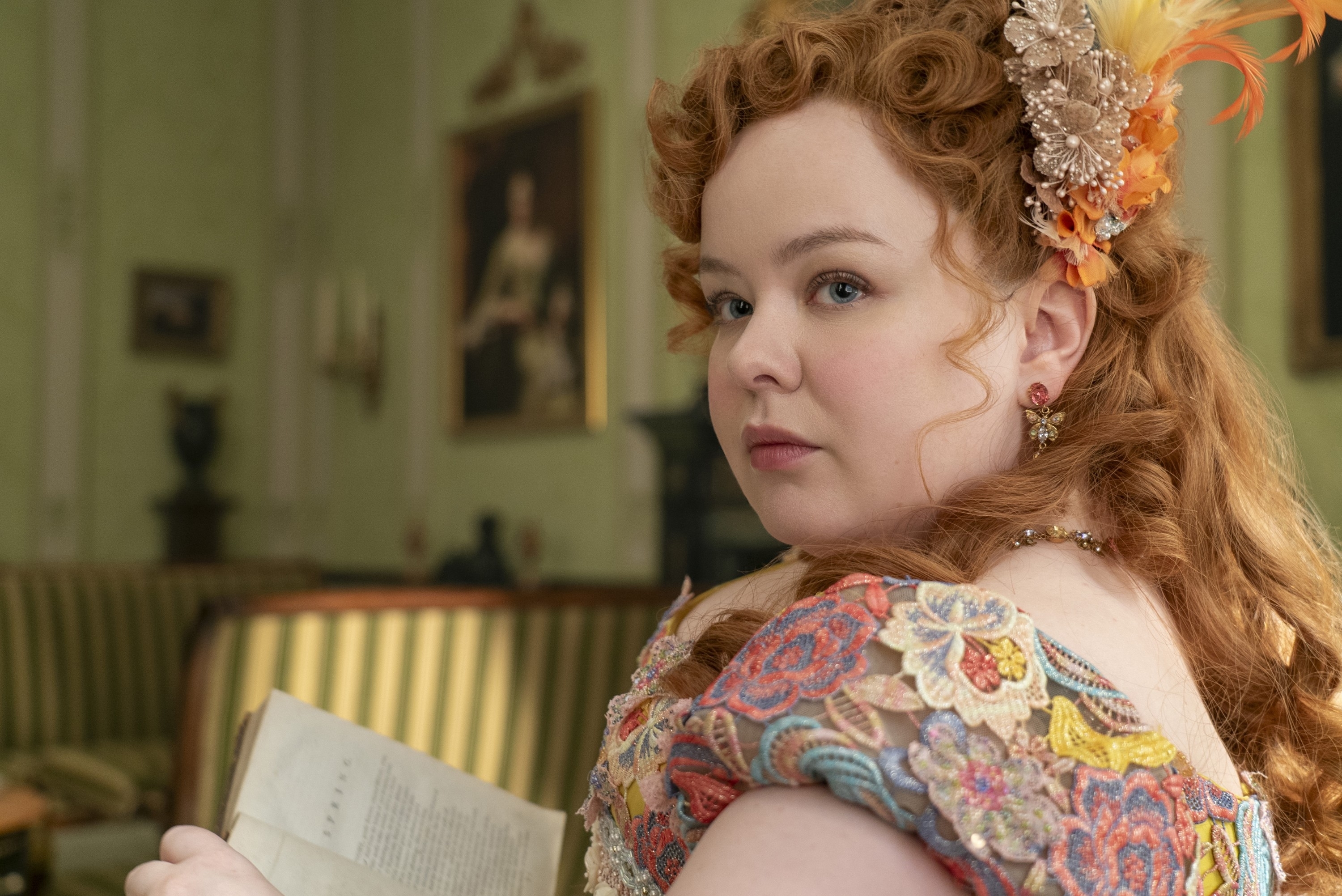 Nicola Coughlan, dressed in a detailed period costume with elaborate floral embroidery and hair accessories, looks over her shoulder while holding an open book
