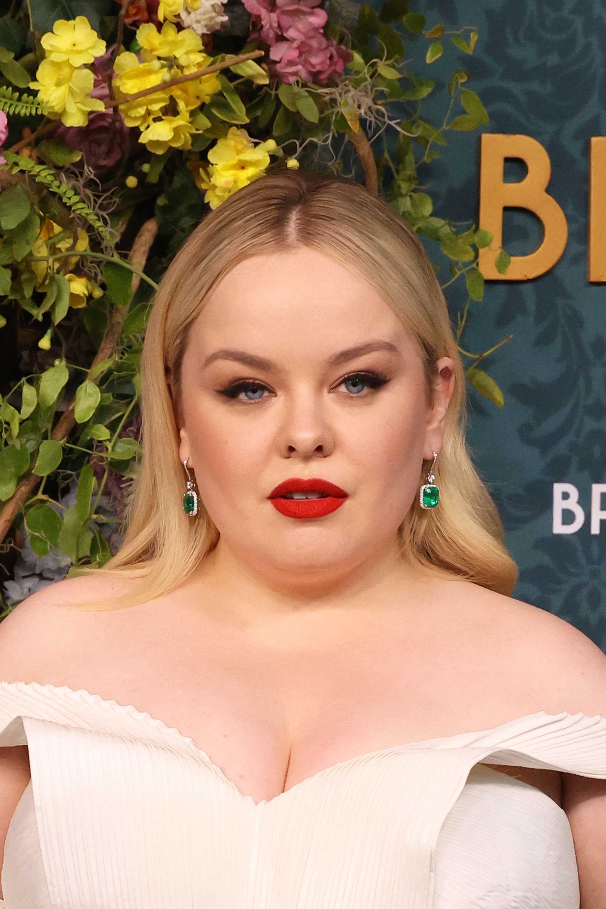 Nicola Coughlan wearing a white off-the-shoulder dress with green emerald earrings, posing in front of a floral backdrop