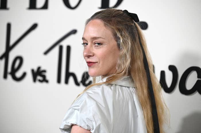 Chloë Sevigny poses in front of a sign reading "Kate vs. the …" wearing a light dress with long sleeves and a black hair ribbon, and looking over her shoulder