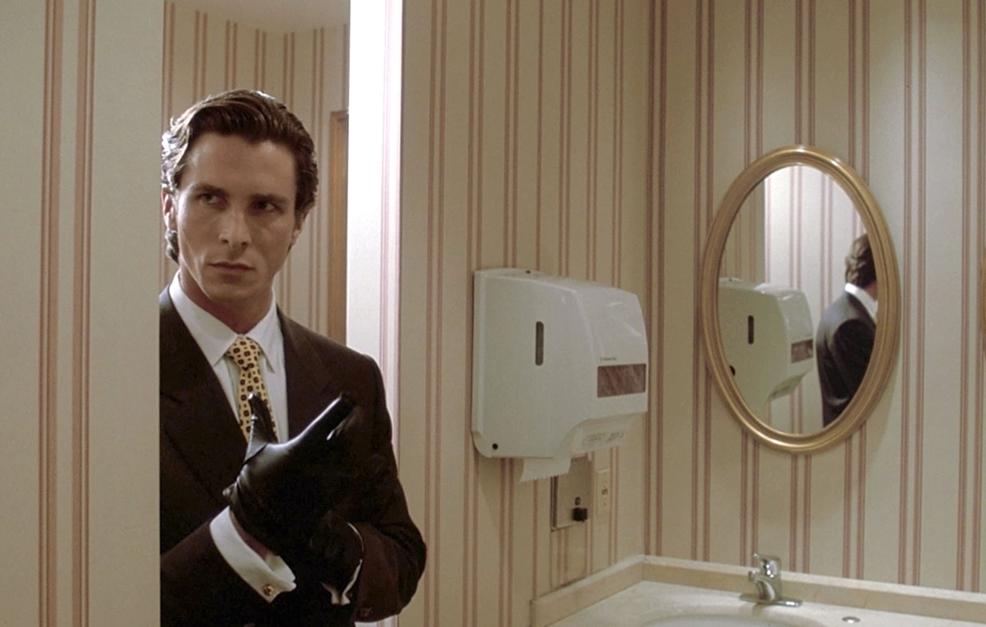 Christian Bale, dressed in a sharp suit with a polka-dotted tie and black gloves, stands in a bathroom, partially hidden behind a door, looking suspicious