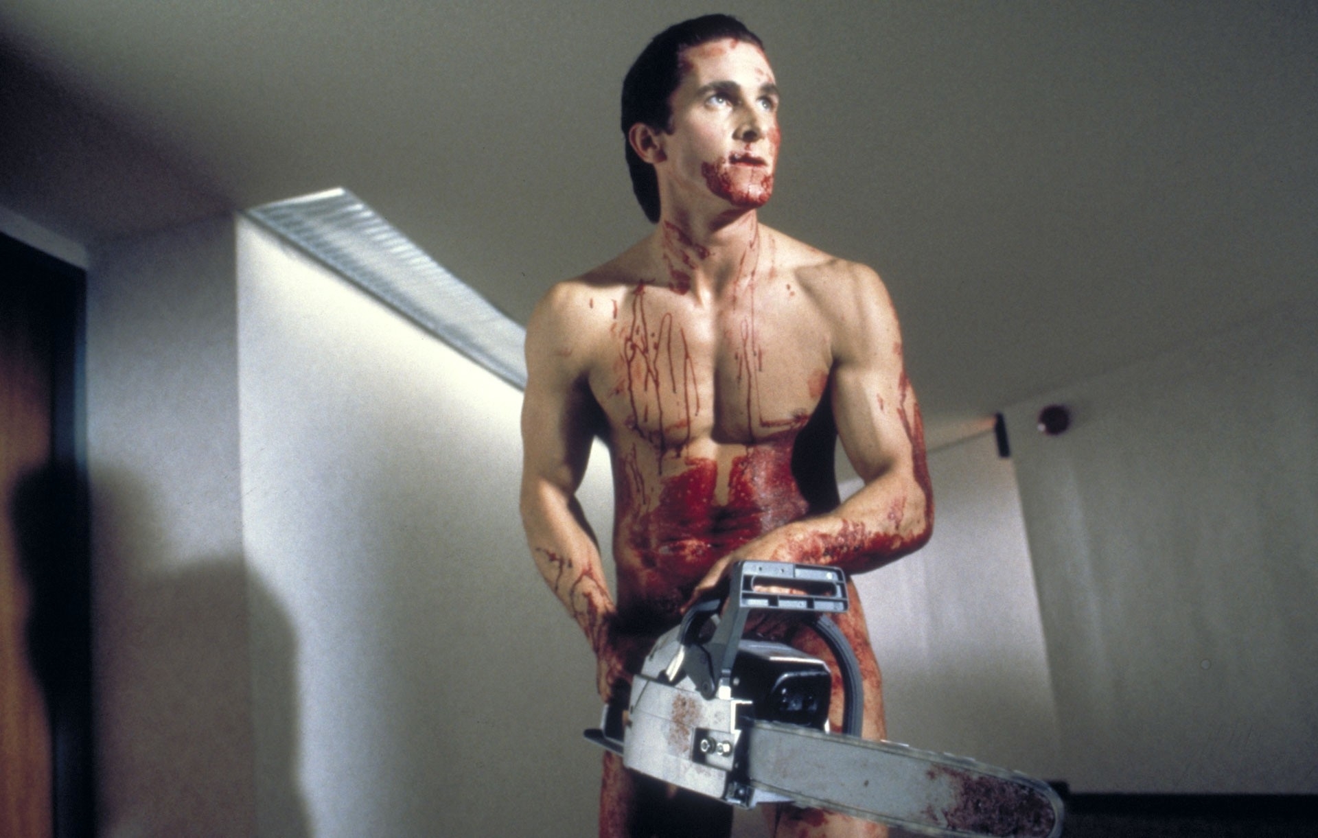 Christian Bale is shirtless, covered in blood, holding a chainsaw, in a scene from the movie "American Psycho."