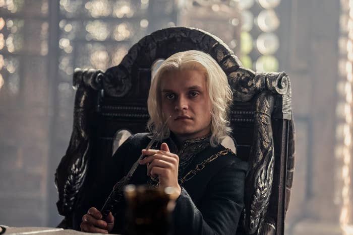 Tom Glynn-Carney as Aegon II Targaryen, seated on an ornate chair, holding a dagger, in a scene from &quot;House of the Dragon.&quot;