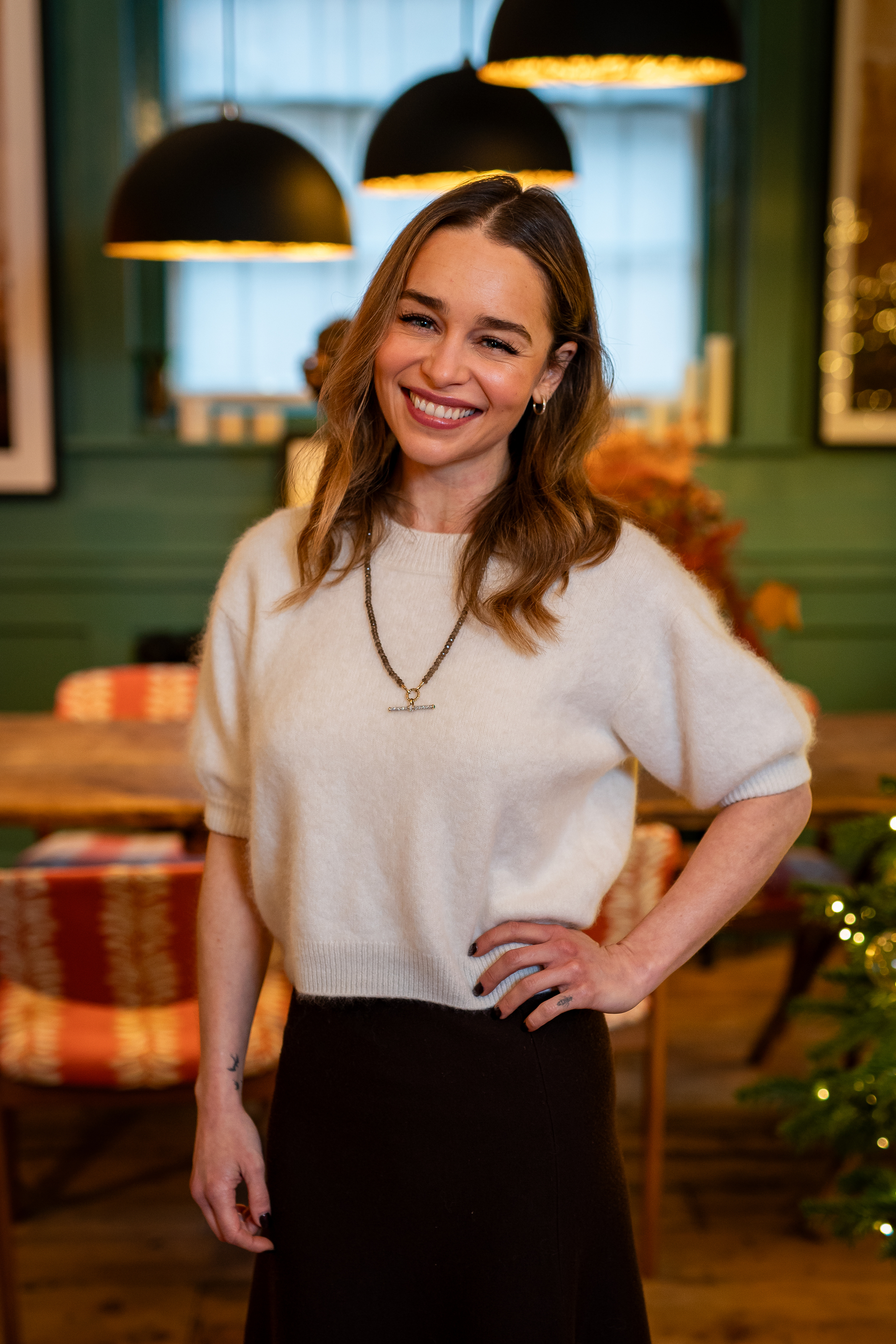 Emilia Clarke smiling indoors, wearing a short-sleeved sweater and a necklace