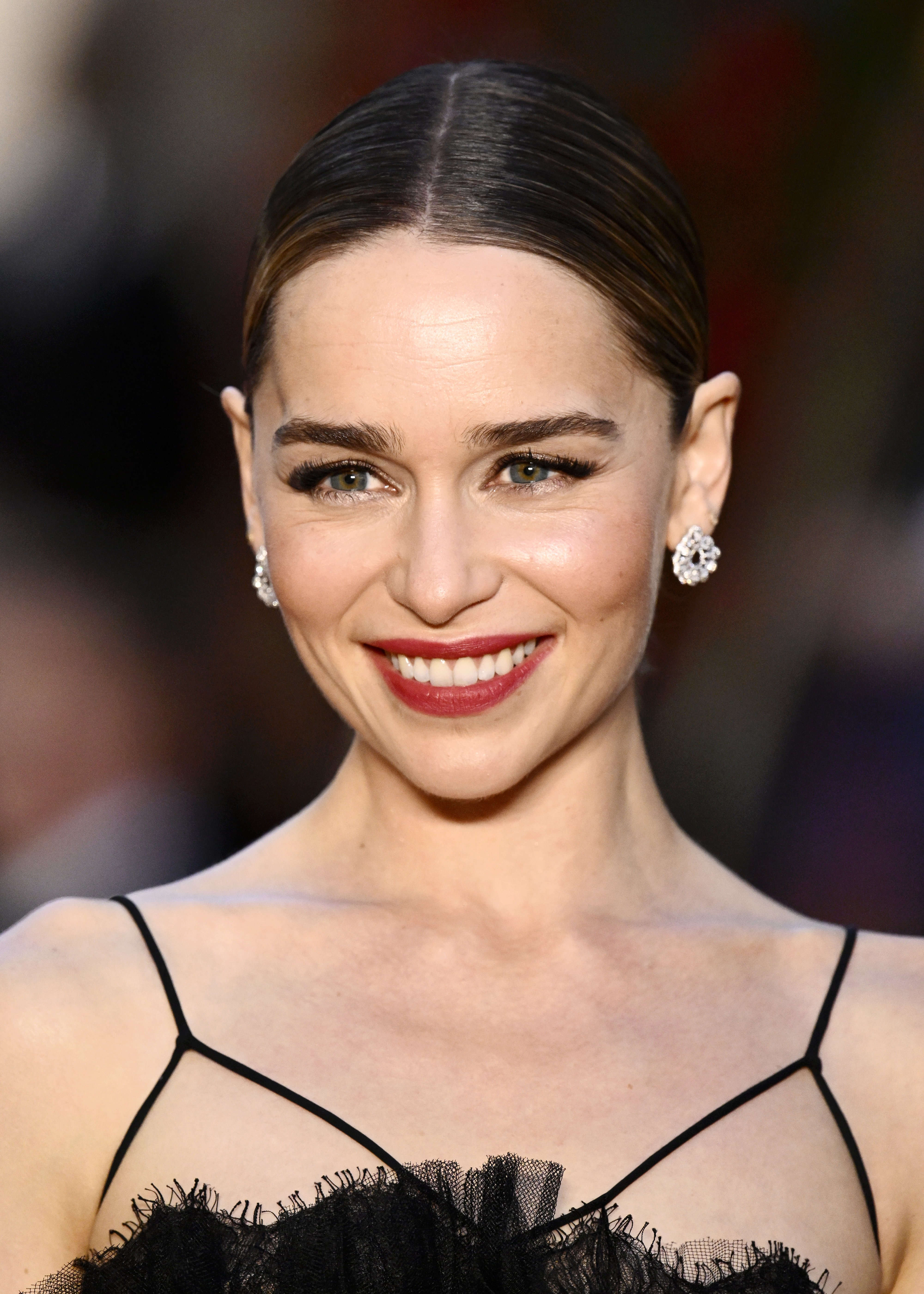 Emilia Clarke smiling, wearing diamond earrings and a black thin-strapped dress with a ruffled neckline. Hair slicked back