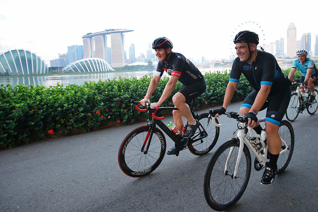 Three people are riding bicycles along a path with a cityscape and waterfront in the background, including the Marina Bay Sands and the Flyer