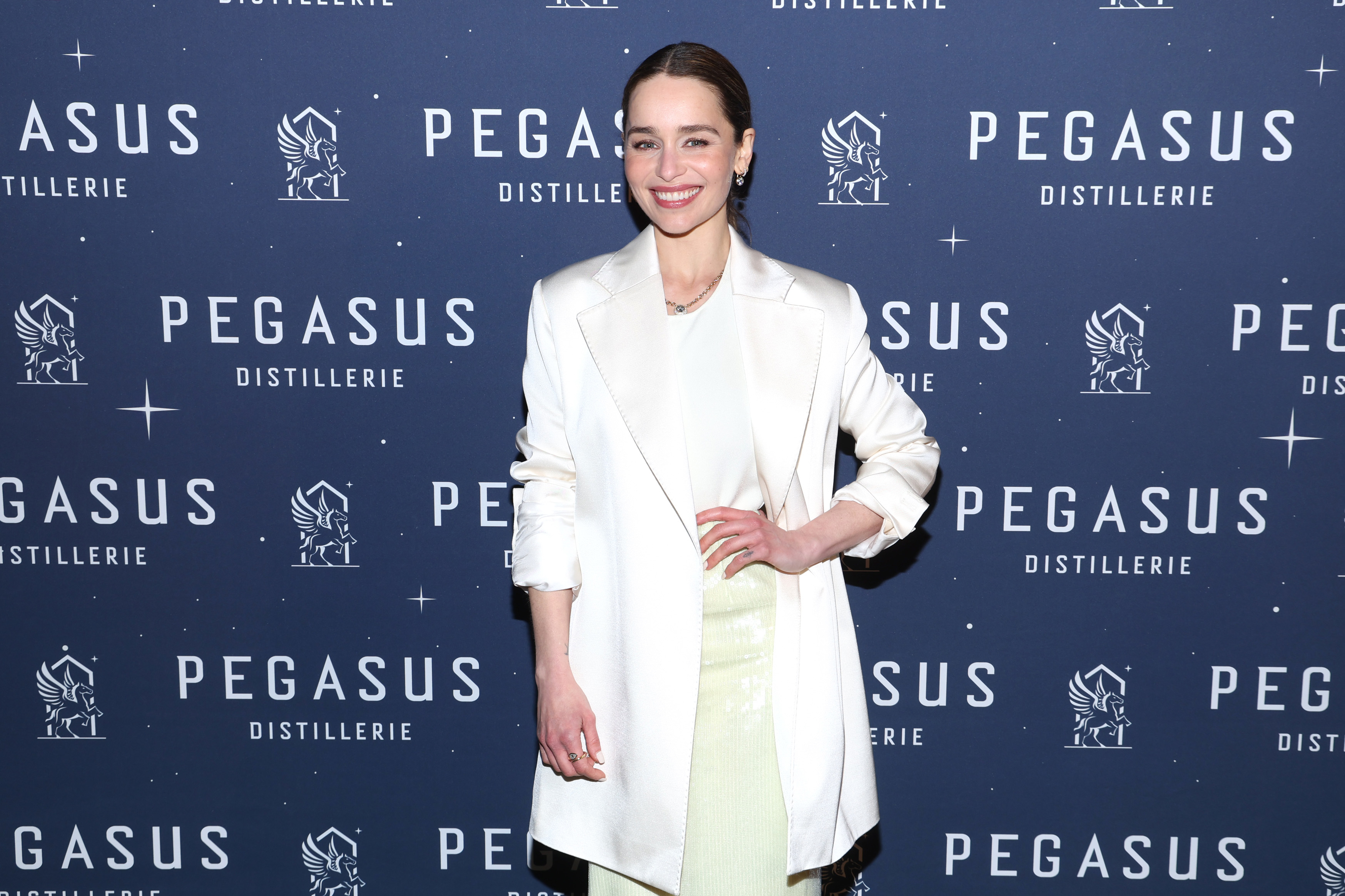 Emilia Clarke is on the red carpet wearing a white blazer over a light dress, posing in front of a Pegasus Distillerie backdrop