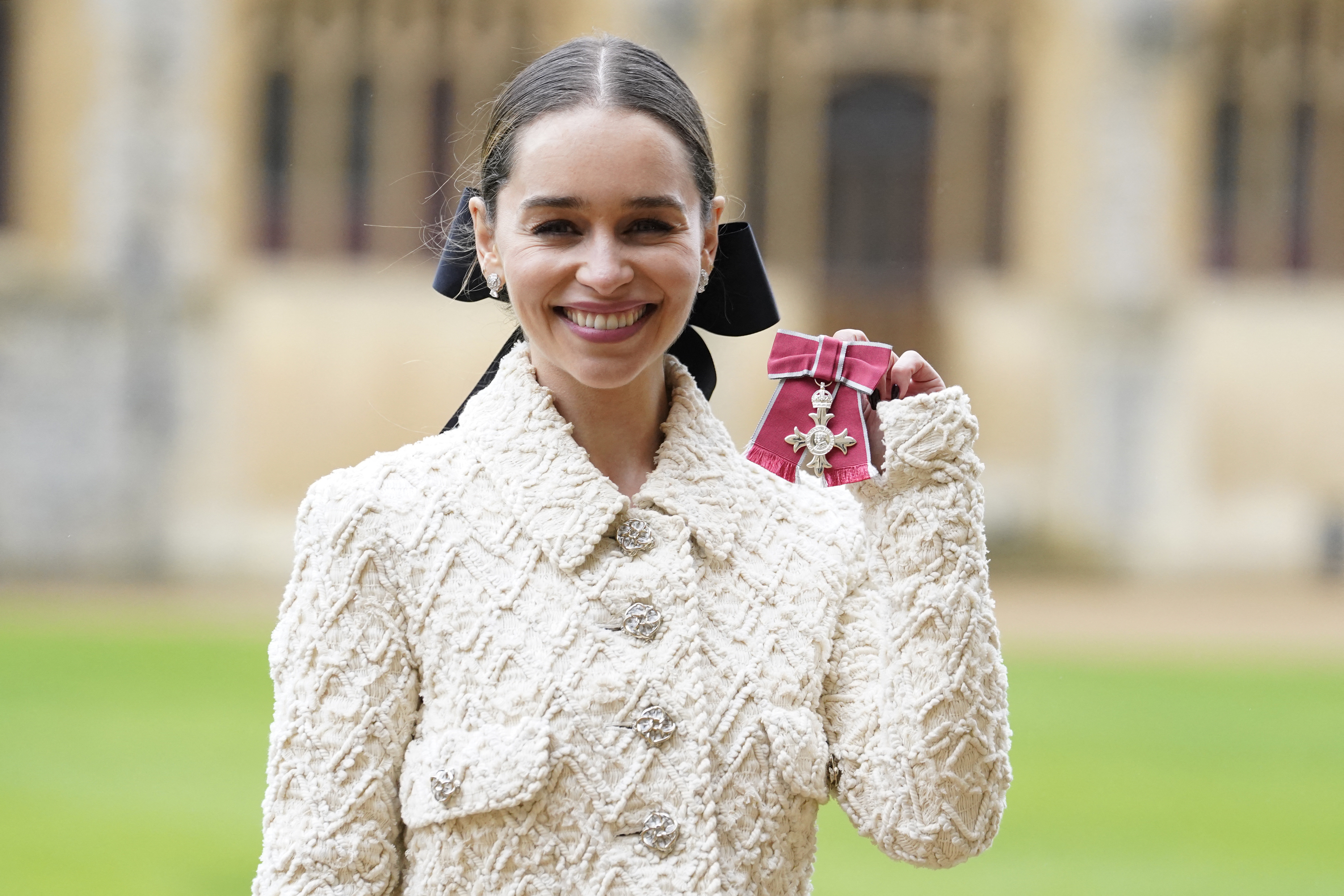 Emilia Clarke smiles and holds her royal honor while wearing an elegant textured coat. She stands in front of a historic building