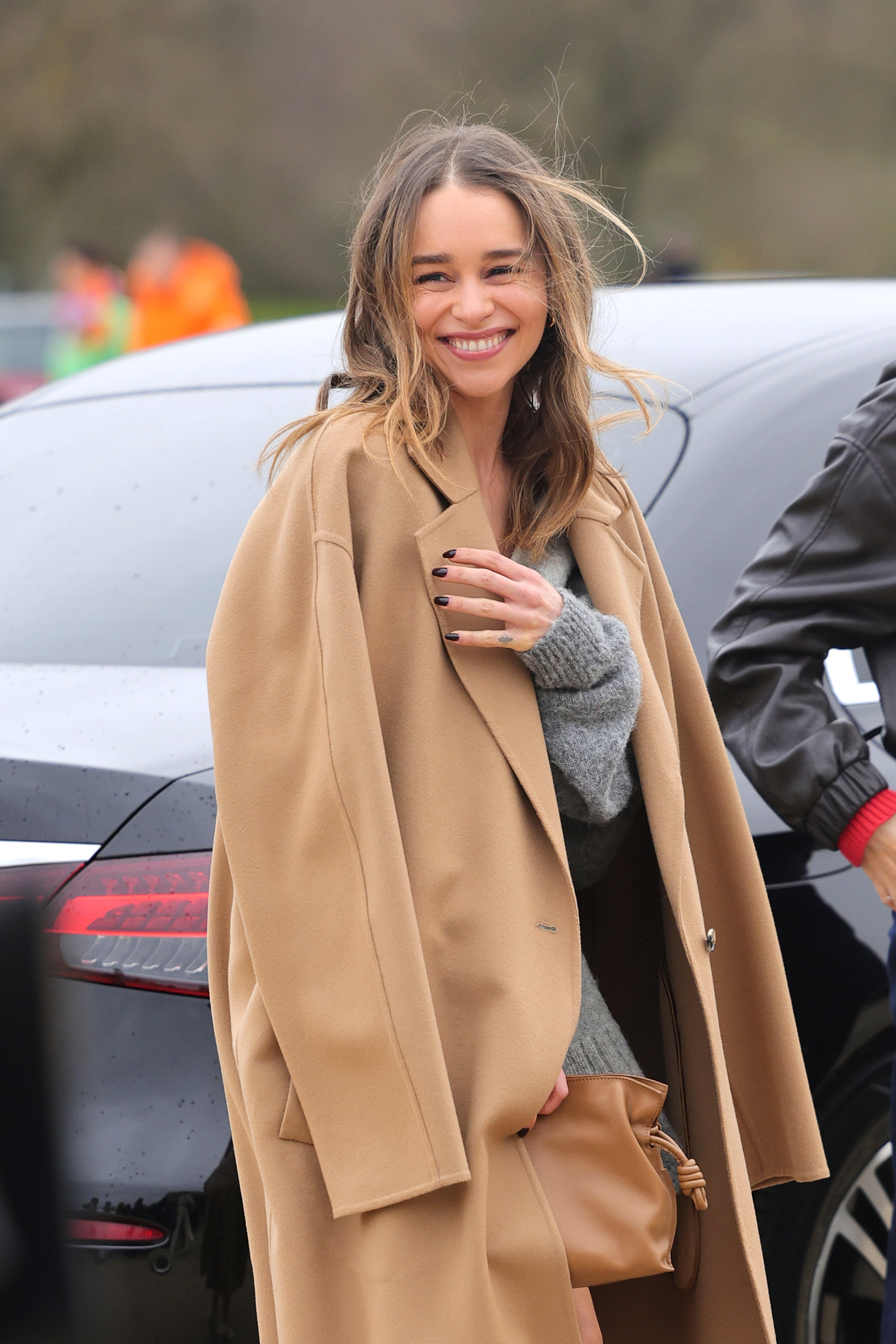 Emilia Clarke smiles while wearing a knee-length coat over a gray sweater and holding a brown clutch bag