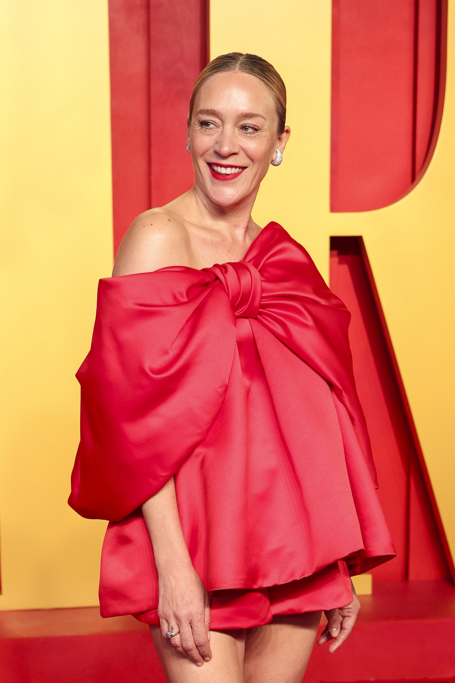 Chloe Sevigny on the red carpet wearing an off-the-shoulder red dress with an oversized bow design, smiling and looking to the side