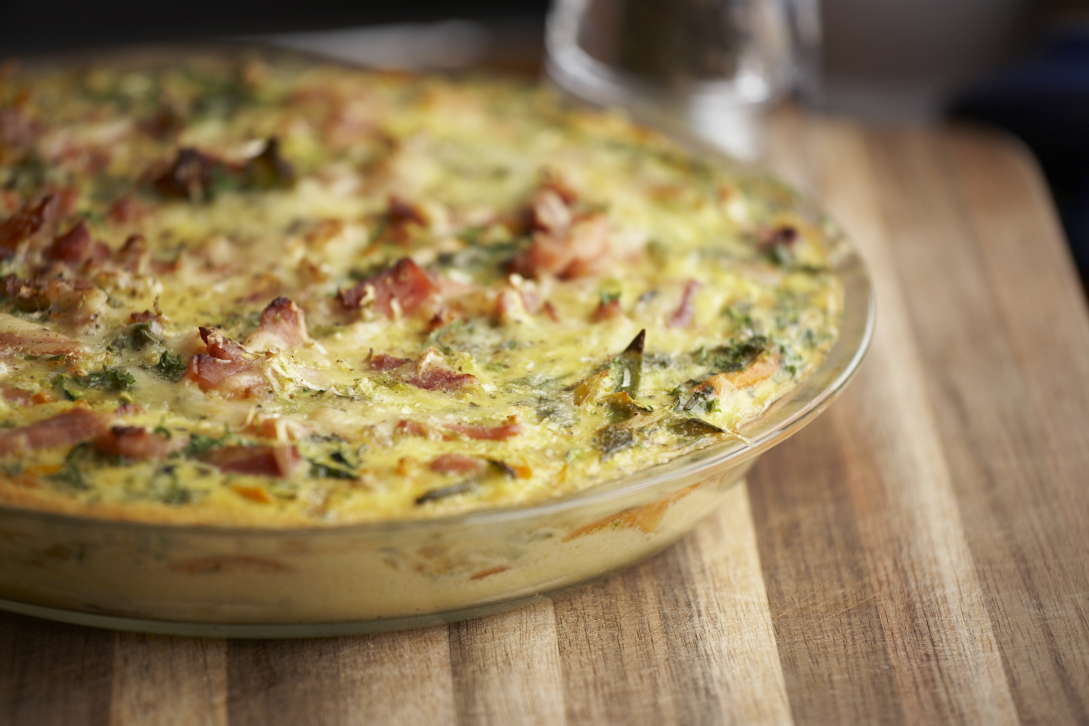 A baked quiche with visible pieces of vegetables and bacon, set on a wooden cutting board