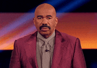 Steve Harvey standing in a suit, looking forward with a surprised expression. Background is a brightly-lit stage setting
