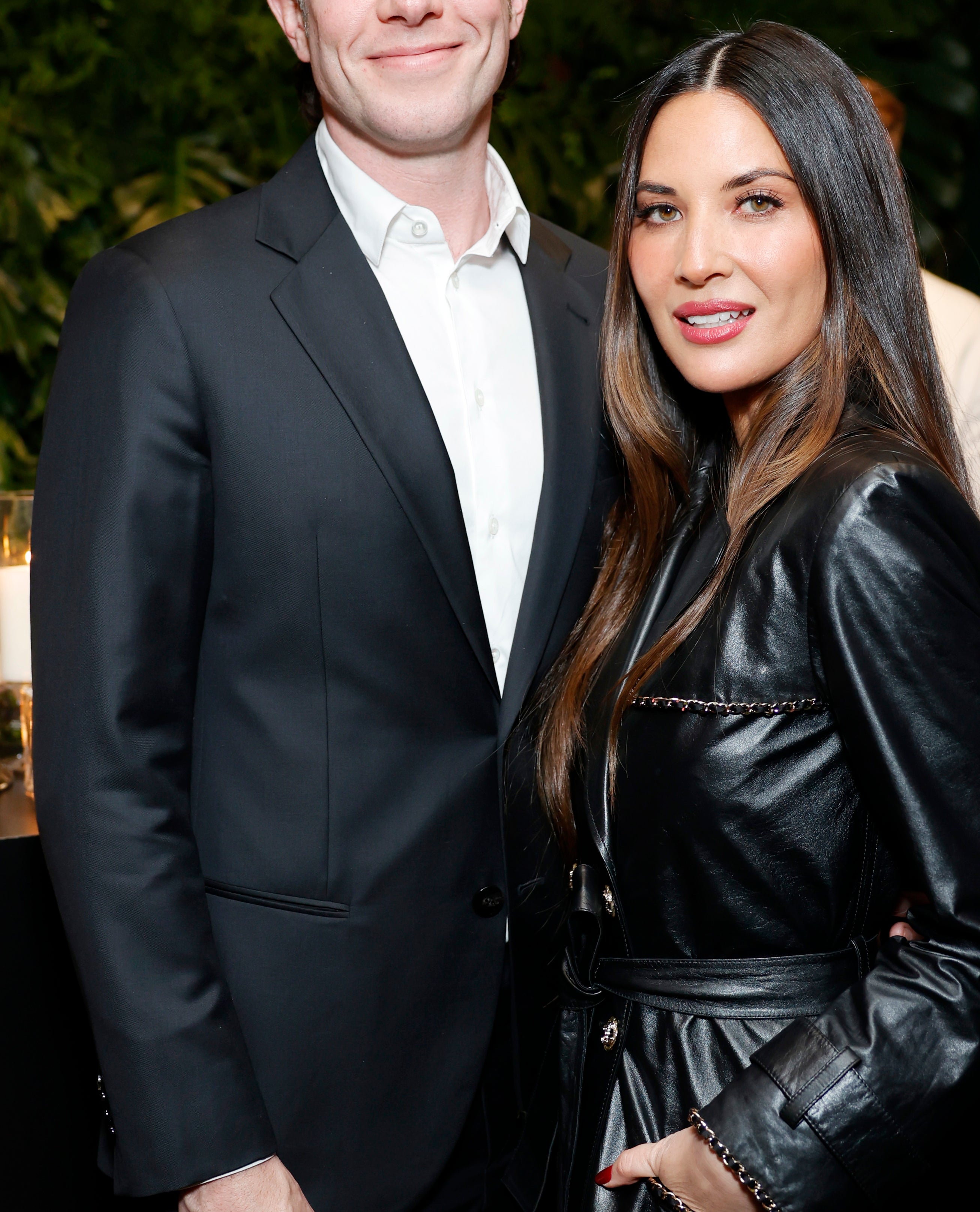 John Mulaney in a suit stands beside Olivia Munn in a leather jacket, both smiling at an indoor event