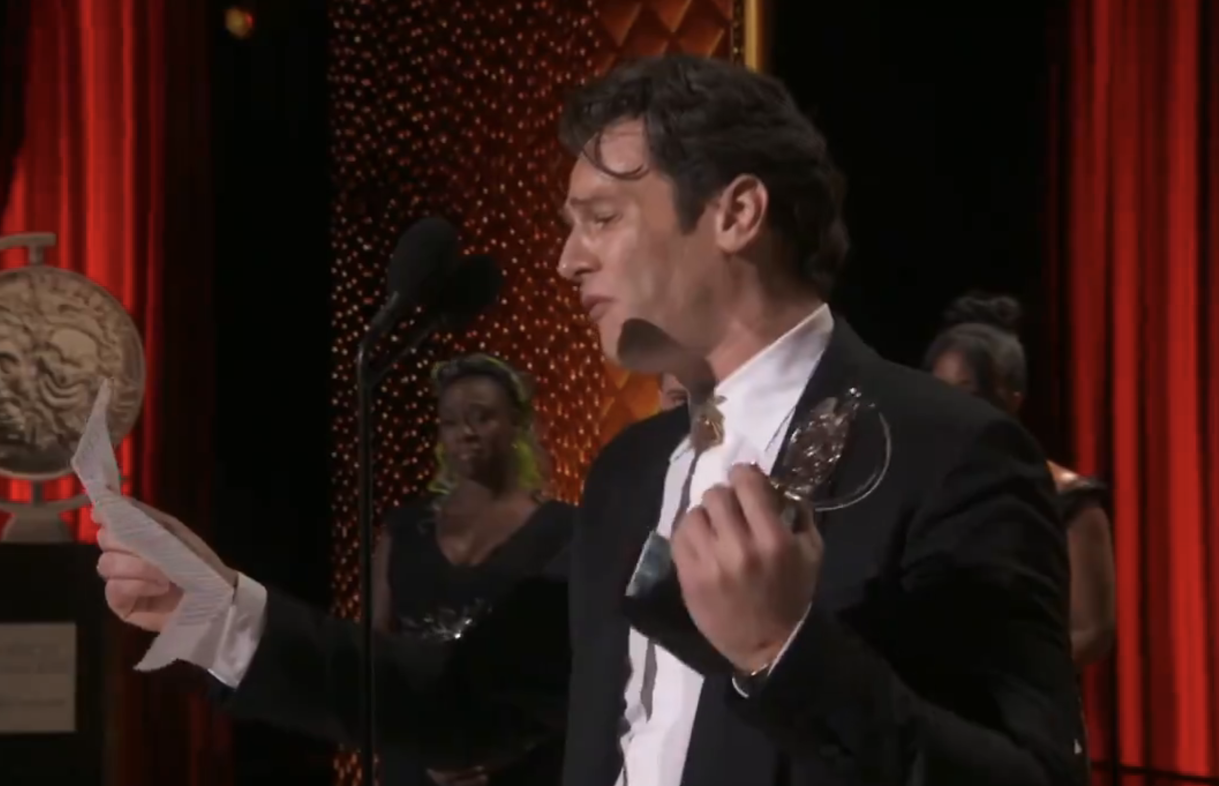 Jonathan Groff, holding a Tony and reading from a piece of paper, gives an acceptance speech on stage, with two people standing in the background