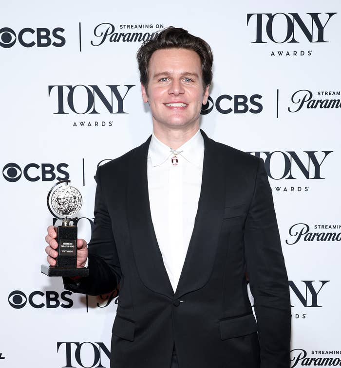 Jonathan Groff in a dark suit and light shirt holding his Tony Award in front of a backdrop with logos for the Tony Awards, CBS, and Paramount+