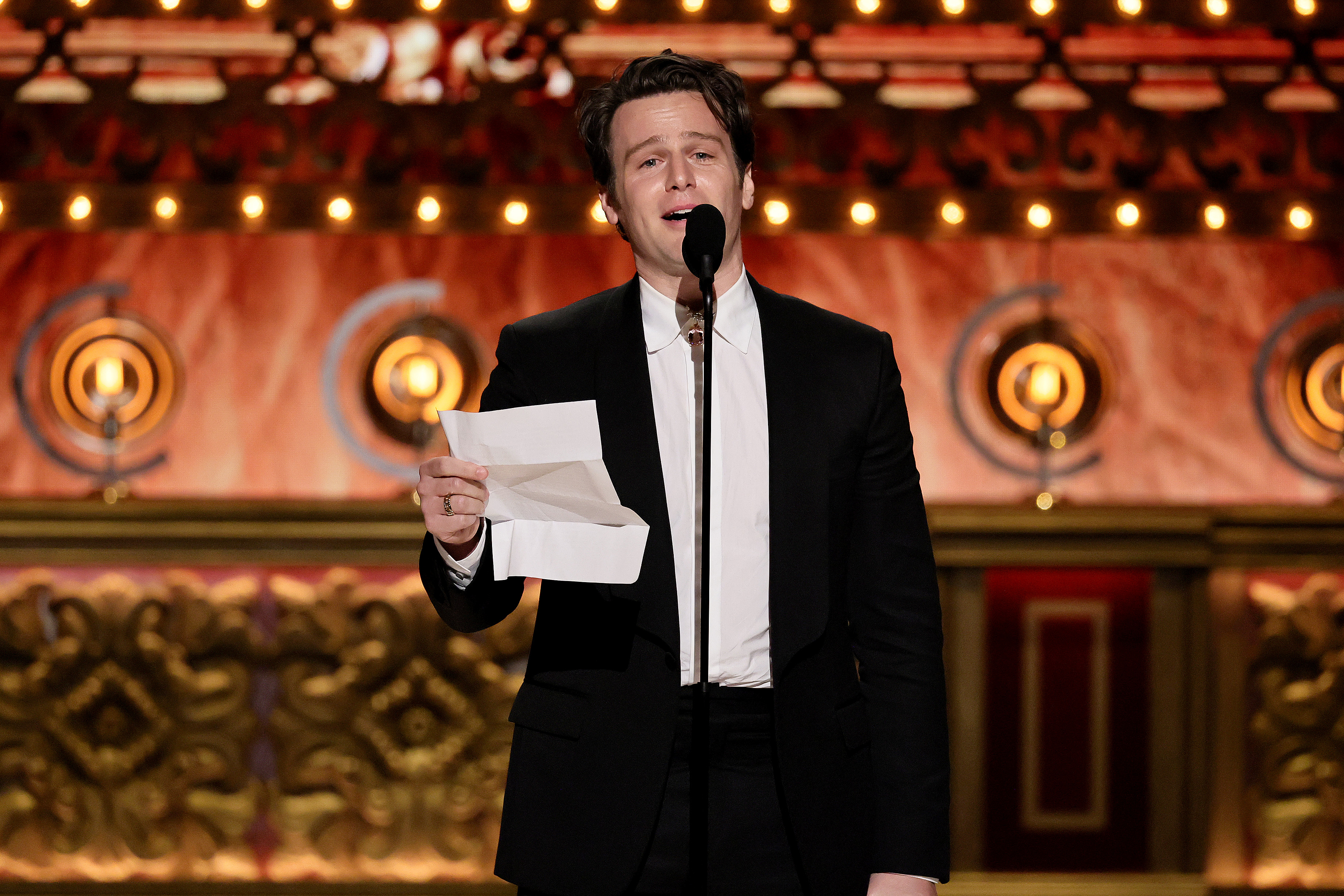 Jonathan Groff speaks into a microphone on stage, holding a piece of paper, dressed in a black suit with a white shirt, at an award ceremony
