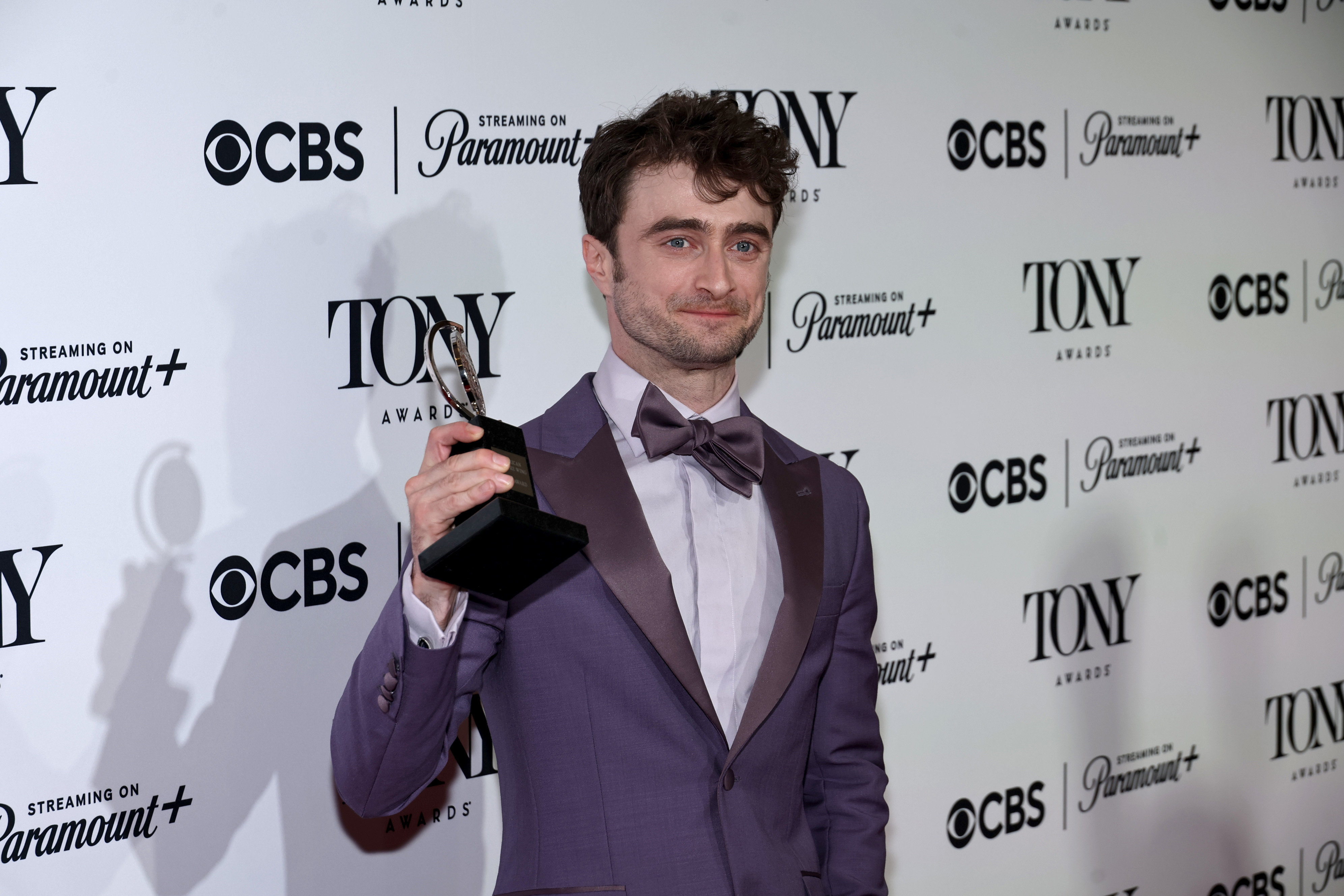 Daniel Radcliffe holding his Tony Award while dressed in a sharp suit with a bowtie at the Tony Awards event backdrop