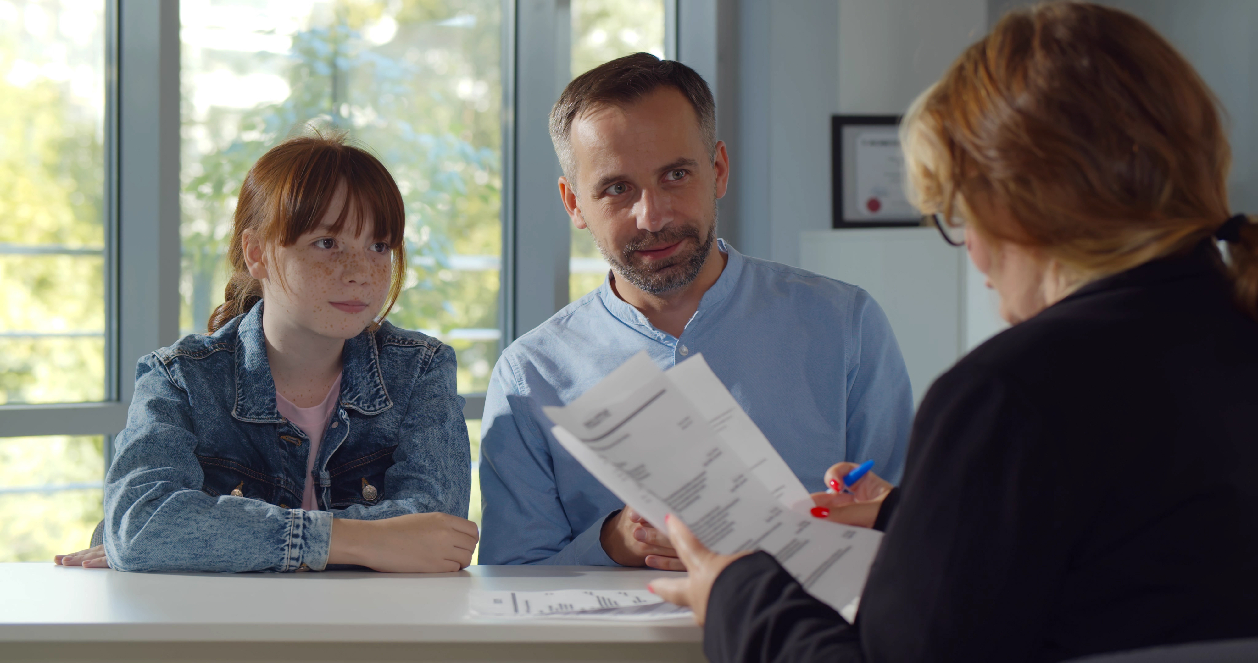 A man and a young girl, wearing casual clothes, sit across a desk from a woman who is reviewing documents. The setting appears to be an office