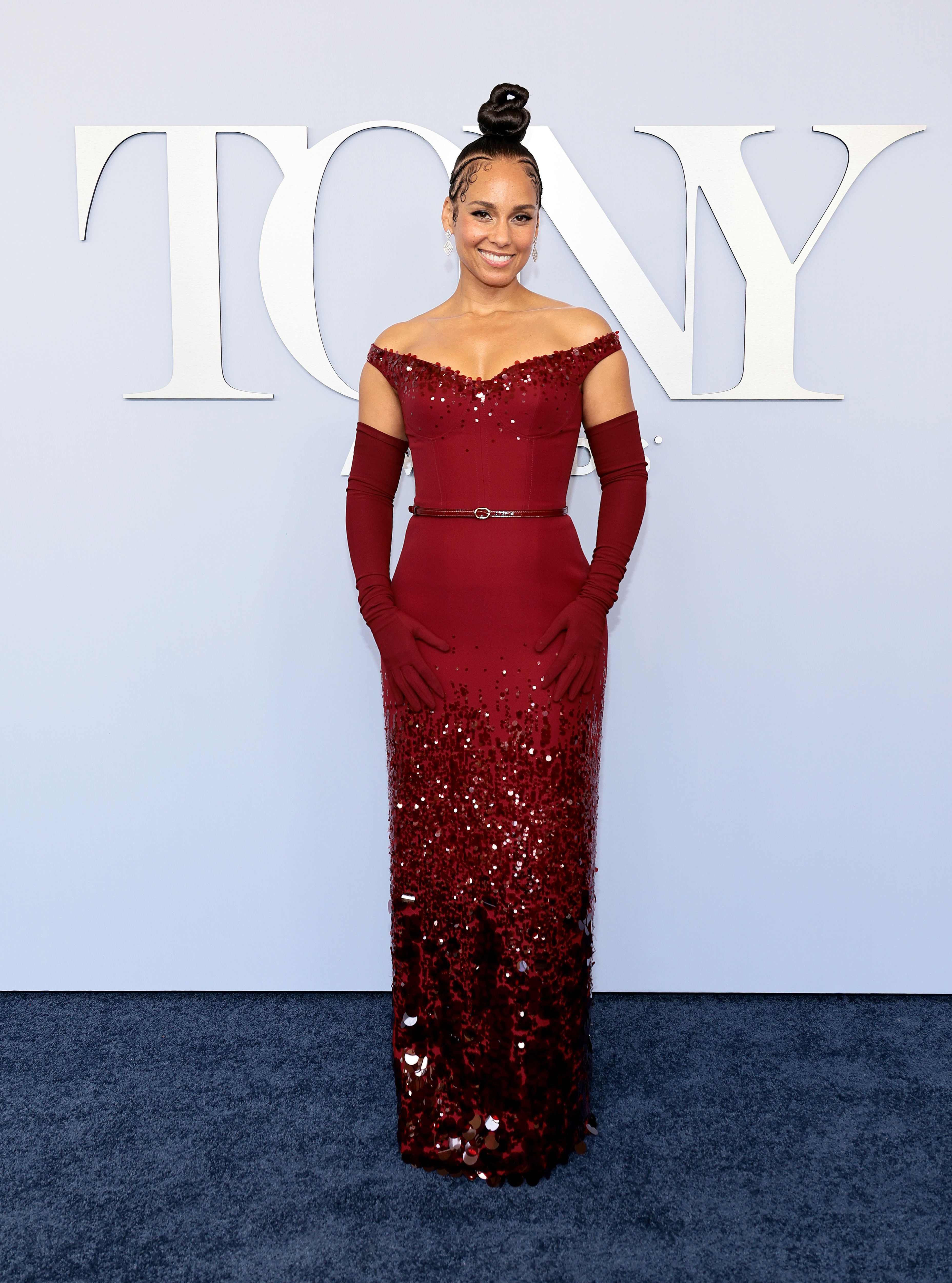 Alicia Keys stands on a red carpet in front of a "Tony Awards" backdrop, wearing an elegant, sequined off-shoulder red gown with matching gloves