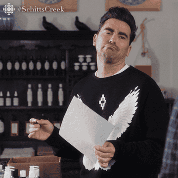 Dan Levy in a scene from Schitt's Creek, smirking while holding a piece of paper with text, wearing a sweatshirt featuring a white eagle
