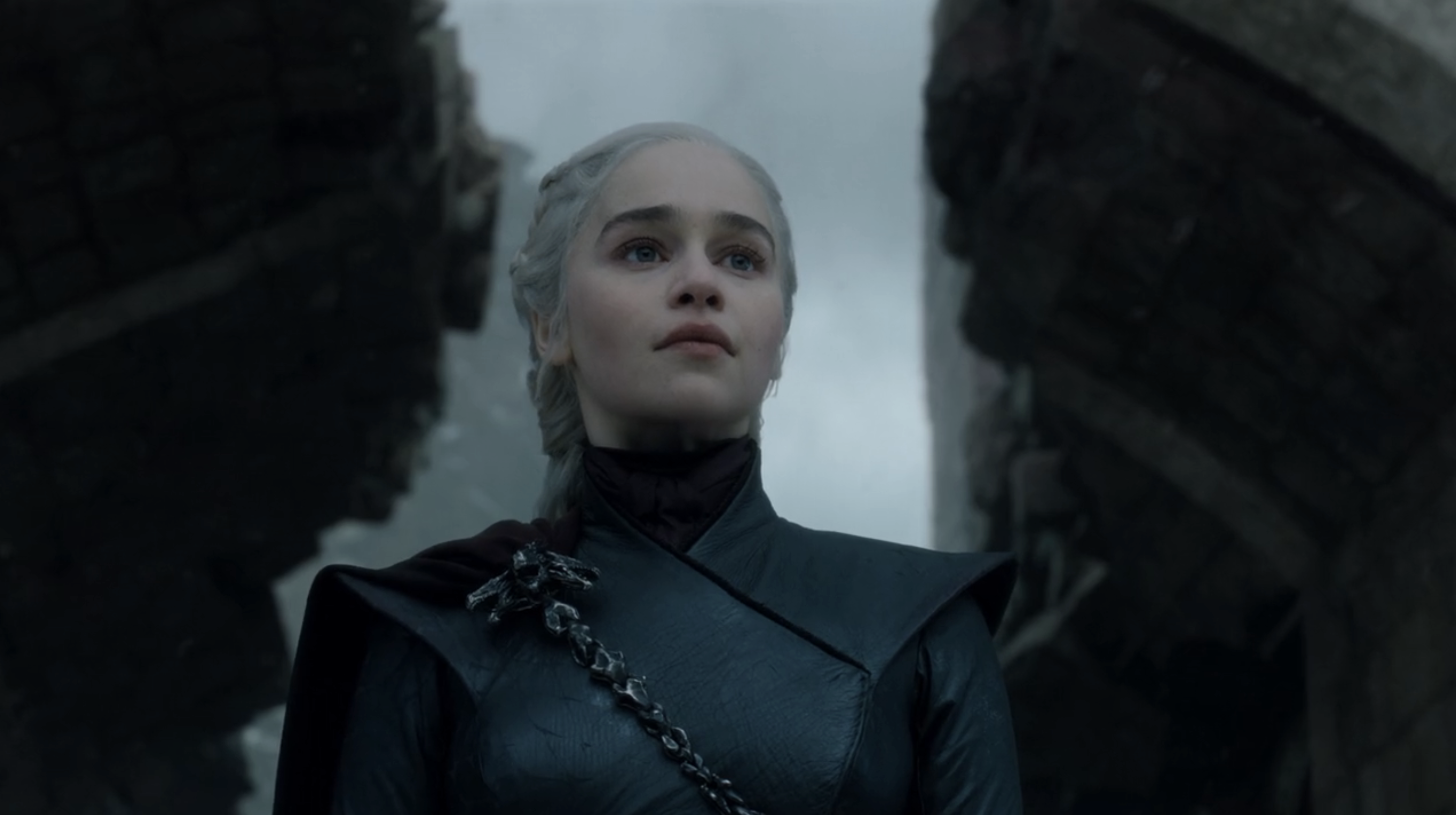 Emilia Clarke as Daenerys Targaryen in a dark outfit from a scene in Game of Thrones, standing between two large, broken structures