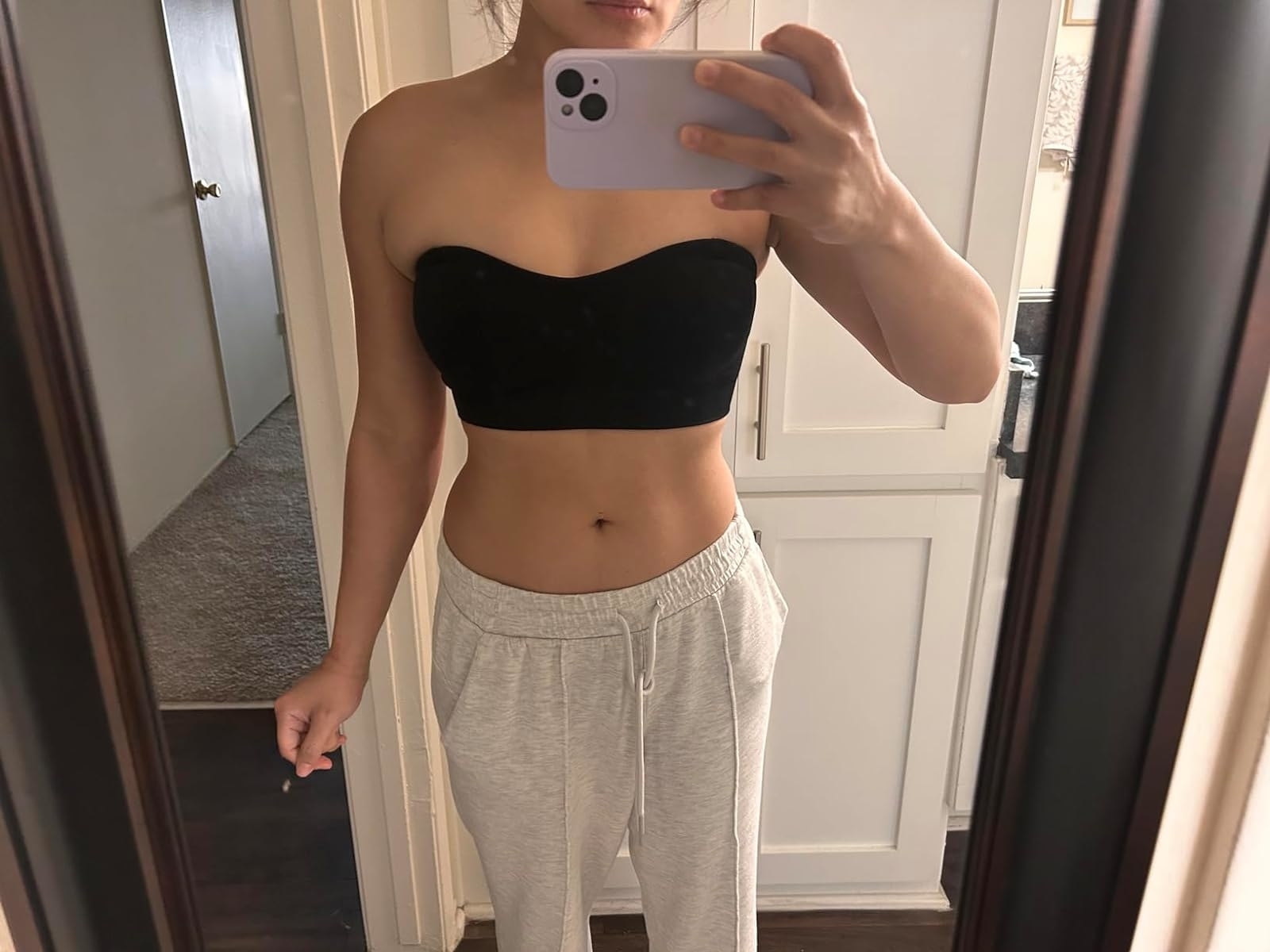 Person wearing a black strapless top and light-colored sweatpants takes a mirror selfie in a modern home