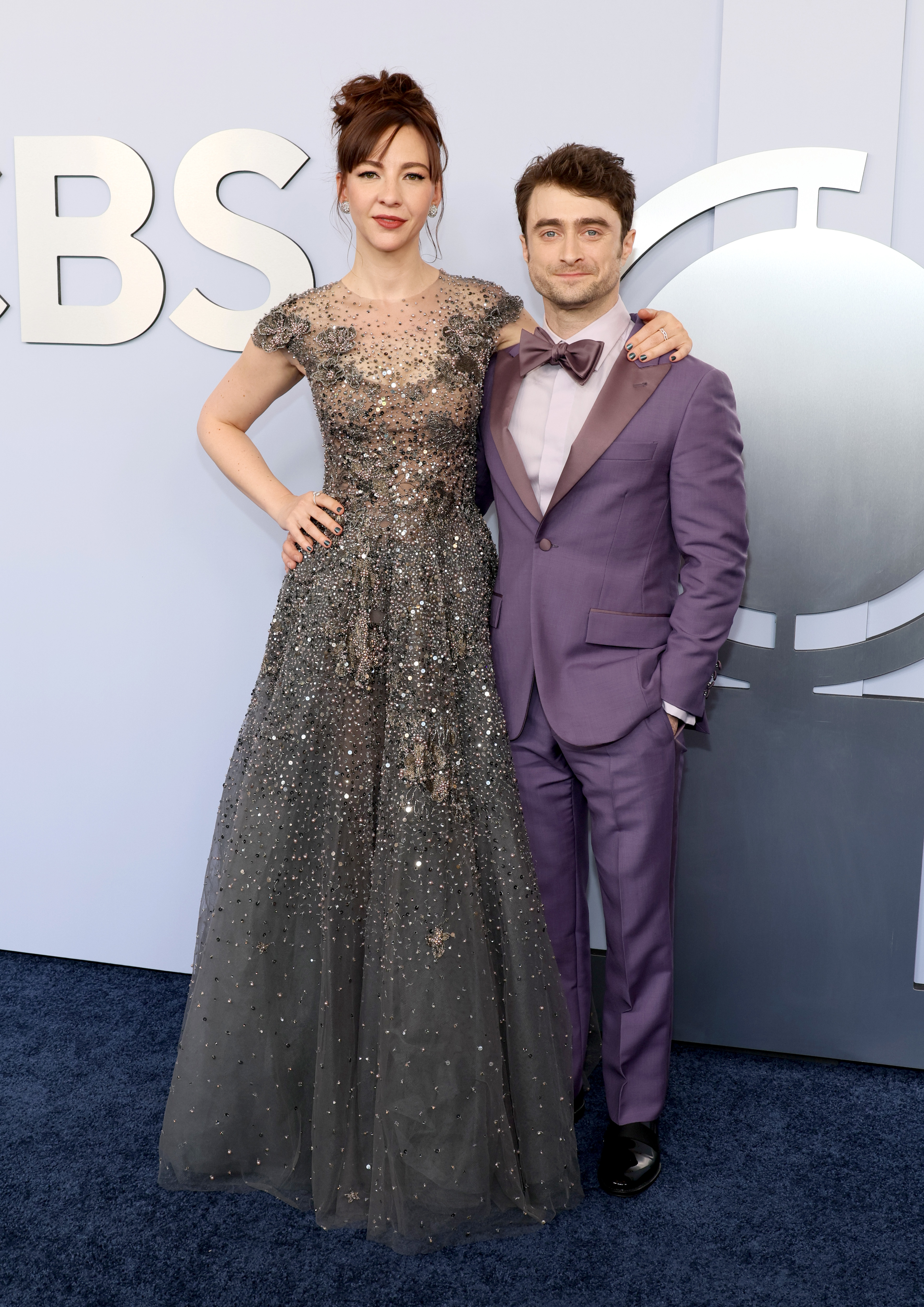 Daniel Radcliffe in a purple tuxedo and Erin Darke in a sparkling, patterned gown pose together on a red carpet at an event