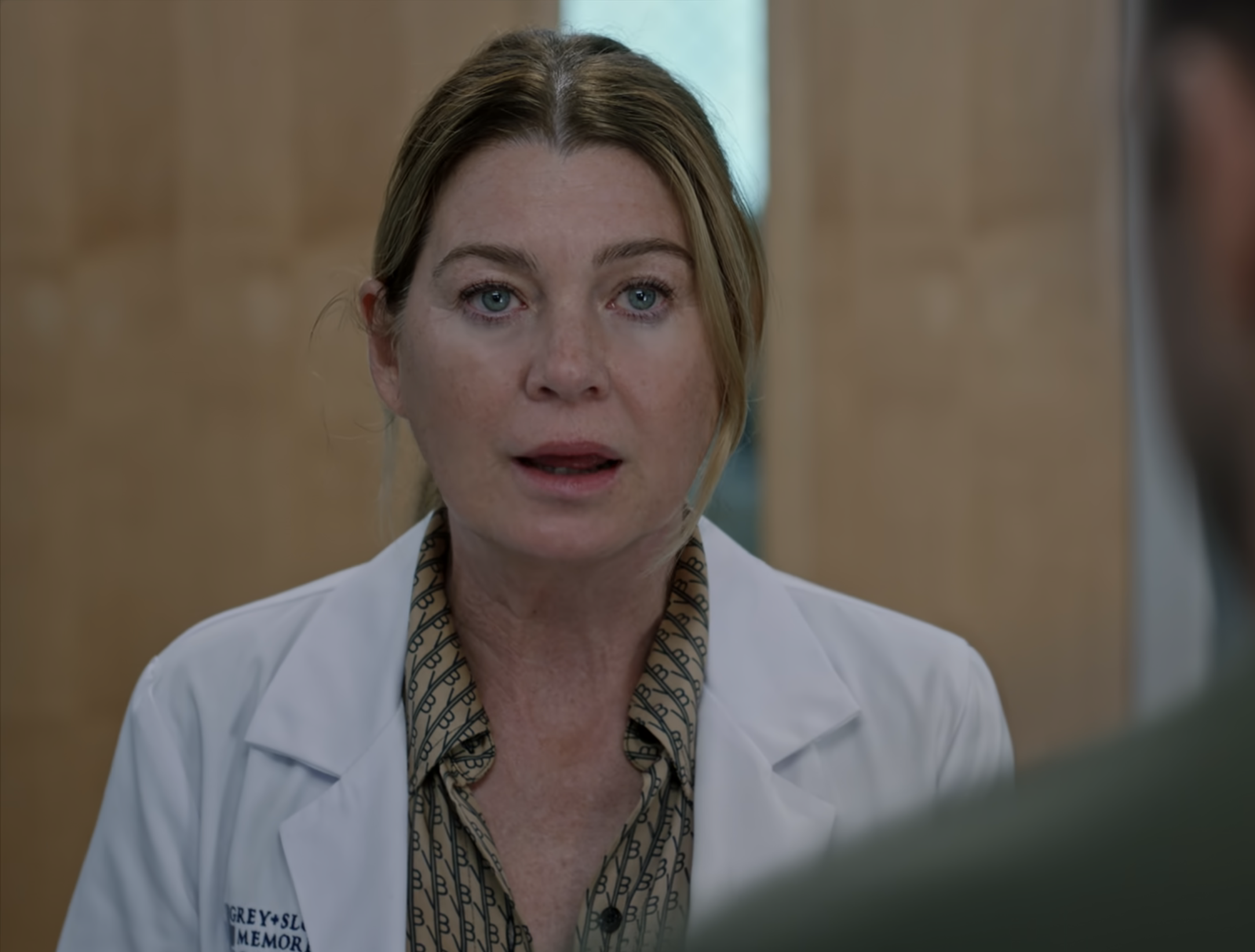 Ellen Pompeo, as Dr. Meredith Grey, speaks to someone off-camera in a scene. She is wearing a doctor's coat over patterned clothing
