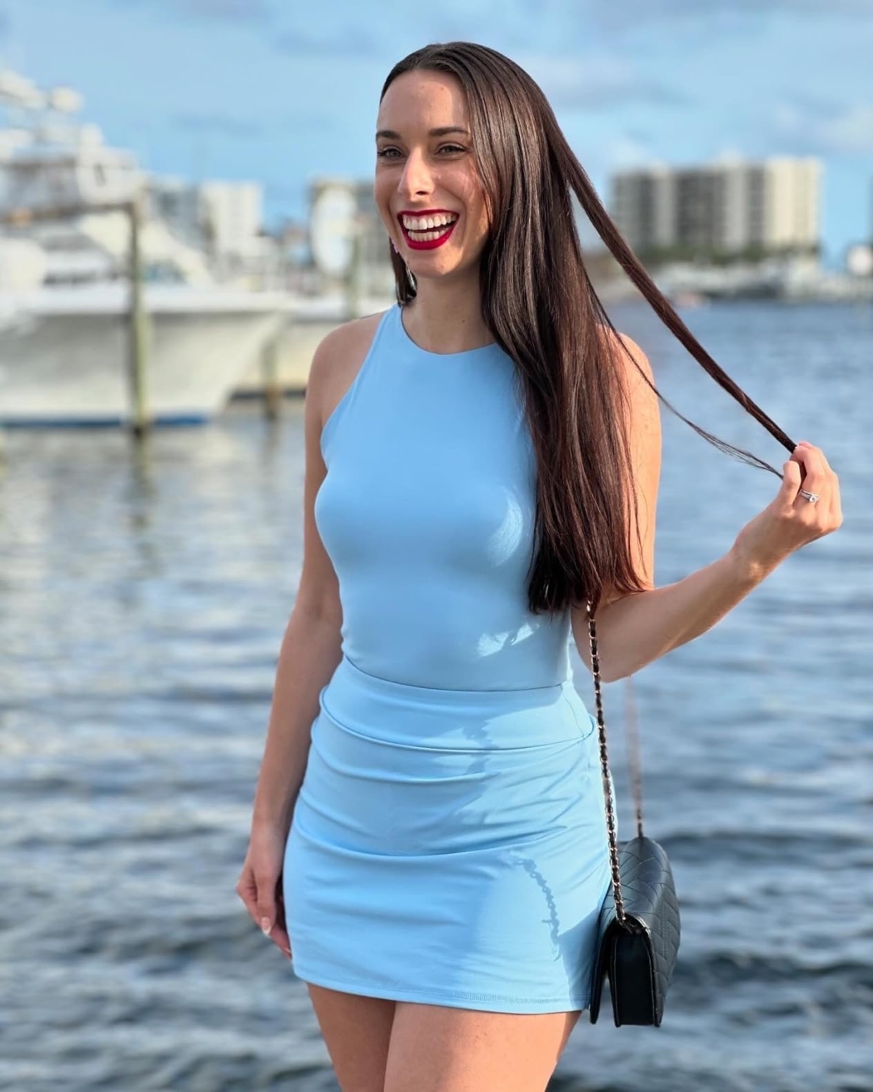 Reviewer with long hair smiles in a sleeveless top and mini skirt by the waterfront, holding a small black purse