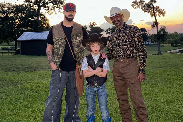 Drake, his son Adonis, and Dennis Graham are standing together outdoors. Drake and Dennis wear cowboy hats, while Adonis is in a cowboy outfit with blue boots