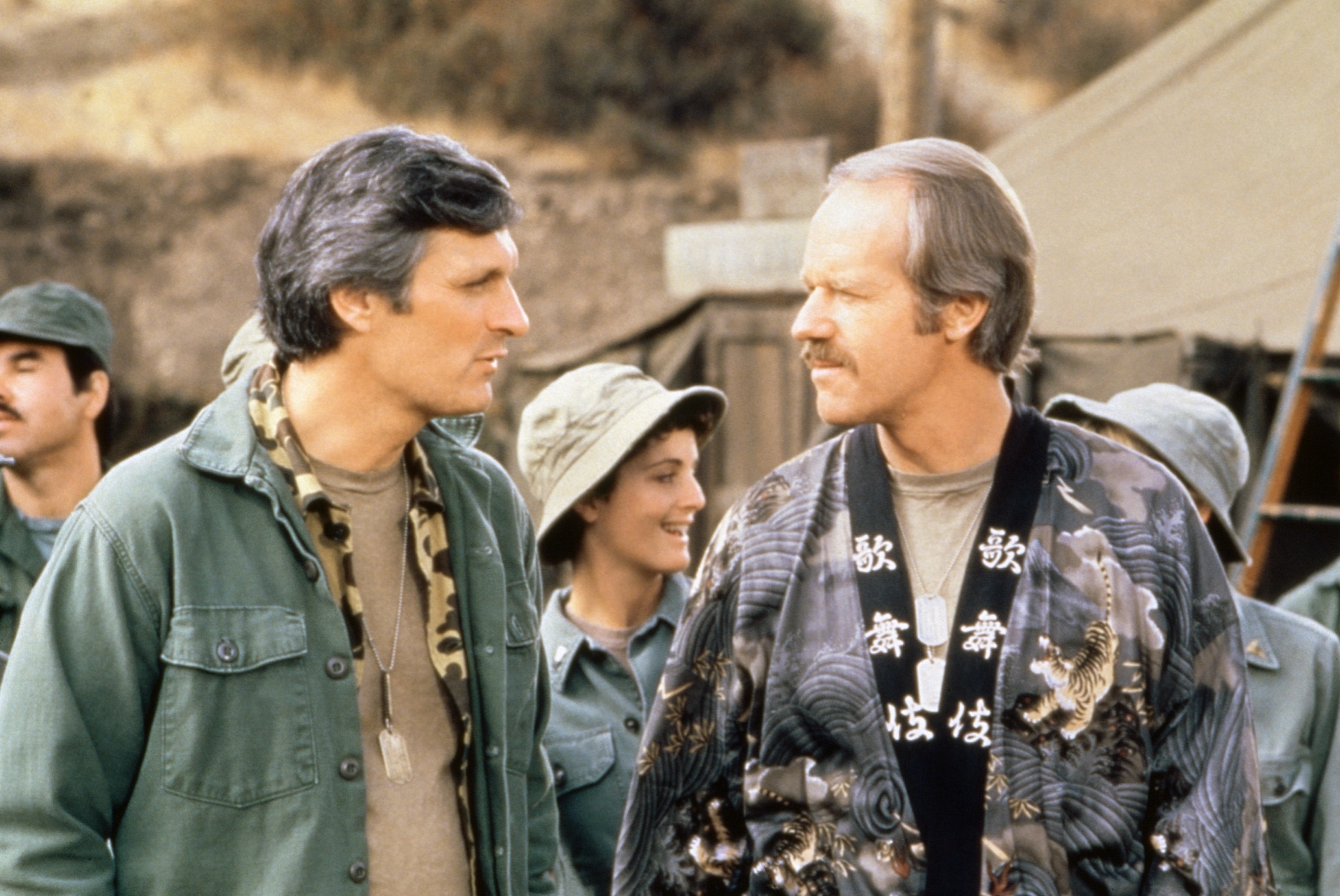 Alan Alda and Mike Farrell in military attire on the set of TV show M<em>A</em>S*H with cast members in the background