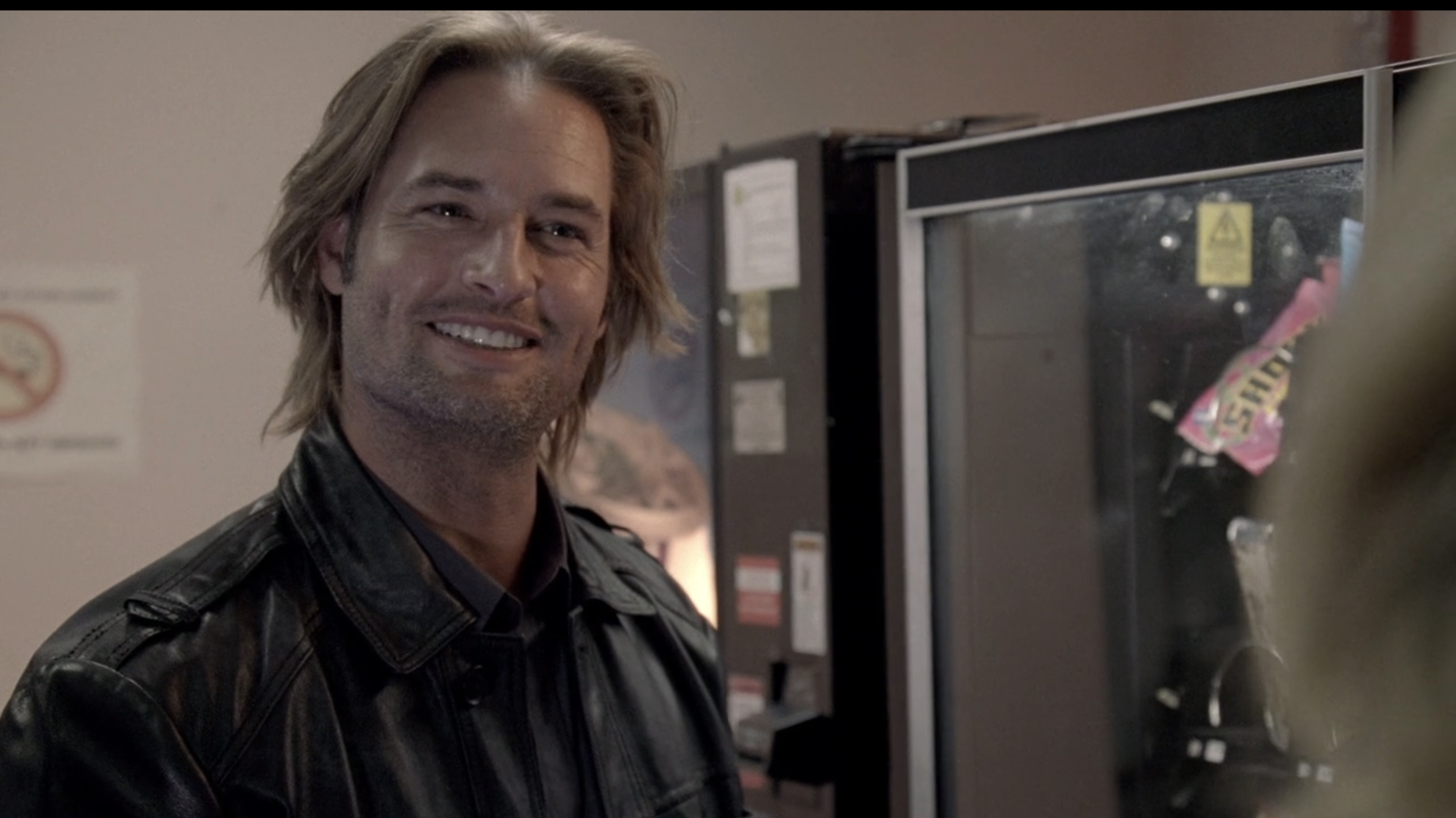 A smiling James Ford with long hair wearing a leather jacket stands in front of a vending machine in a dimly lit room