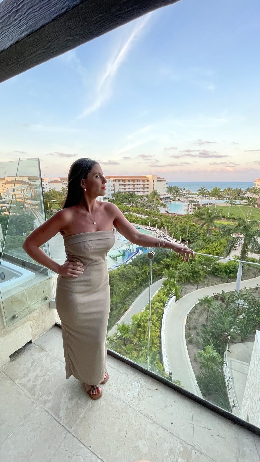 A reviewer dressed in a form-fitting strapless dress stands on a balcony overlooking a lush resort with a pool and buildings in the distance